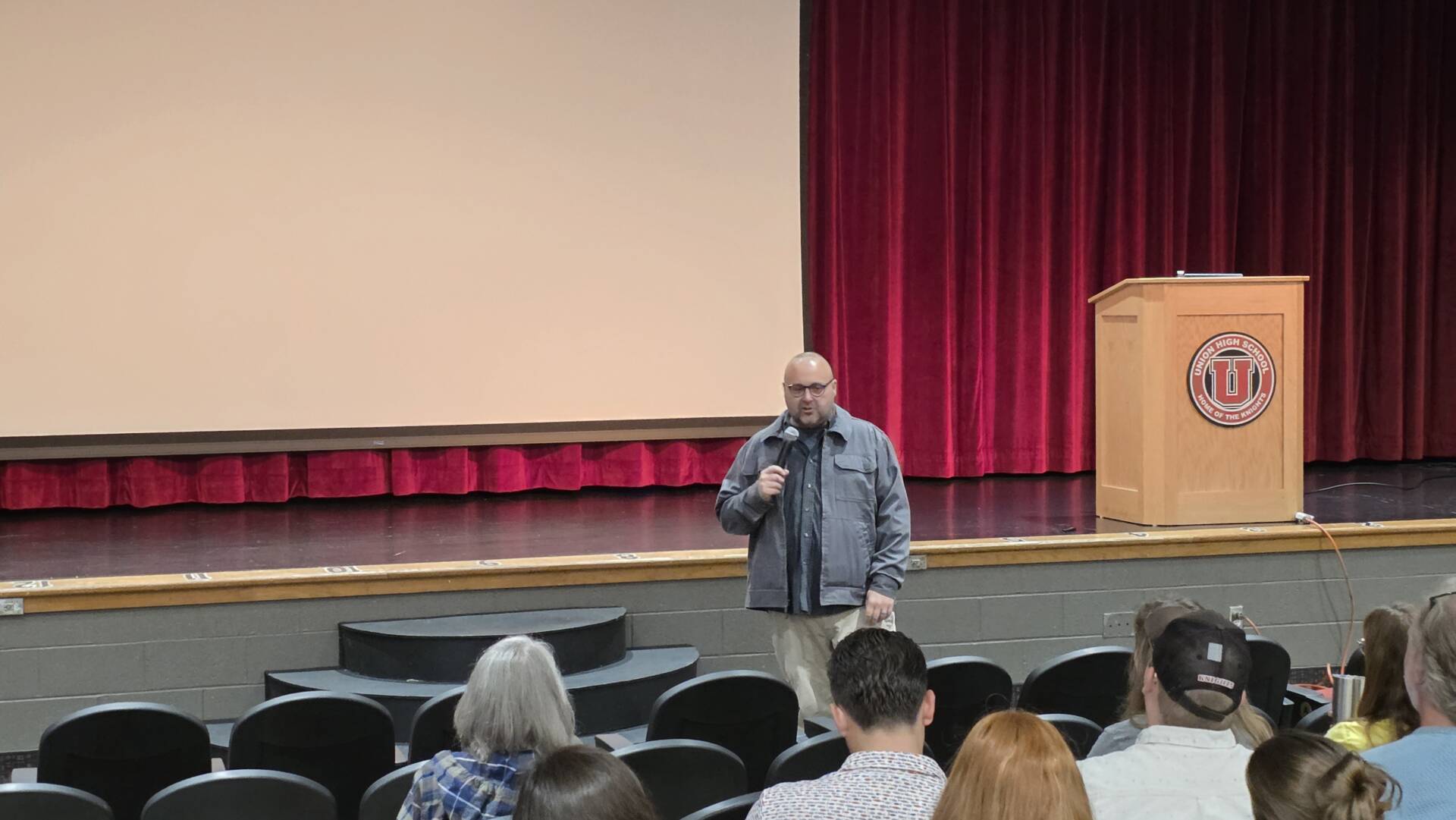 A man holding a microphone addresses a seated audience in a school auditorium with red stage curtains.