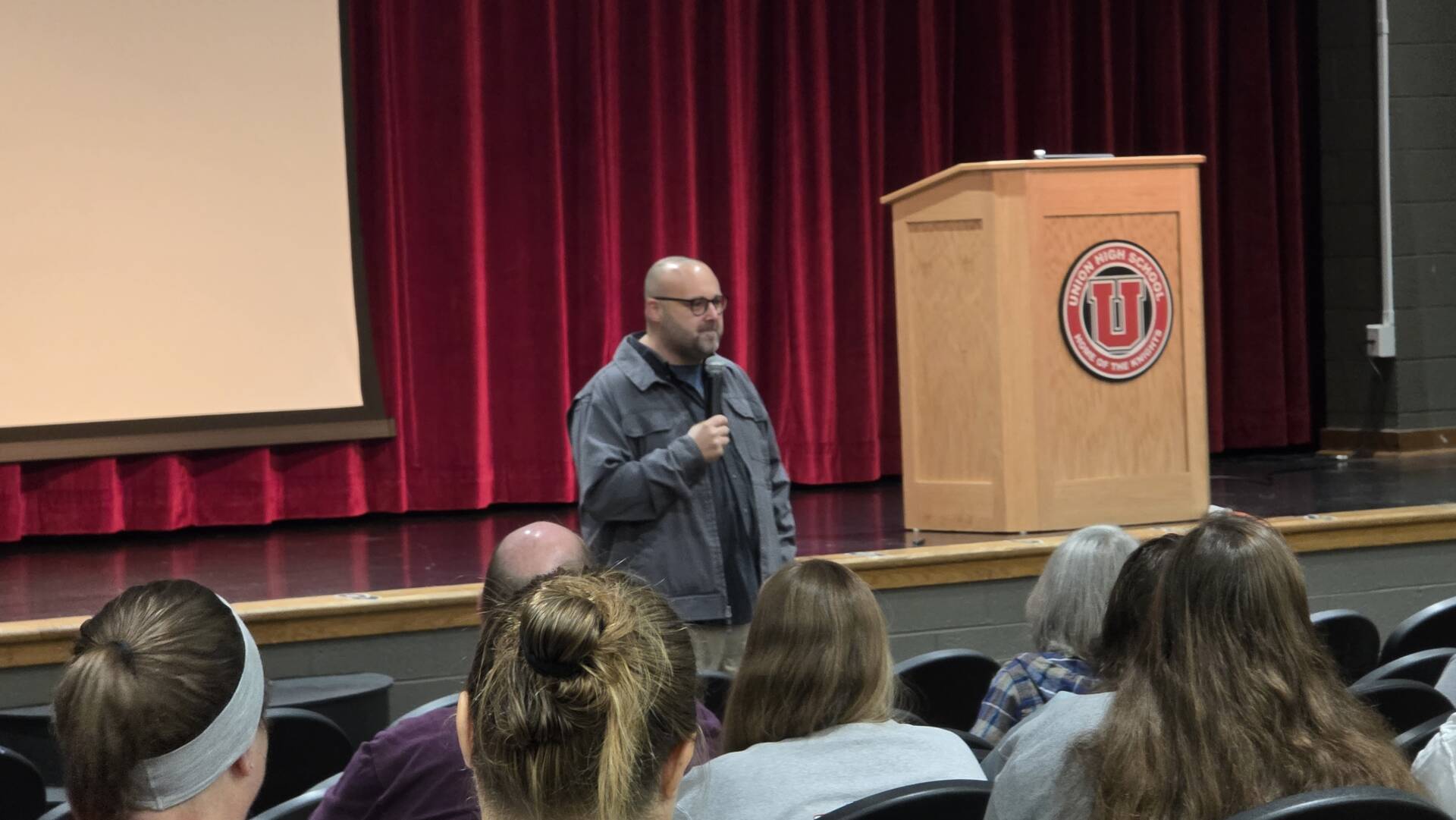 A man with a microphone speaks to an audience in front of a red-curtained school auditorium stage.