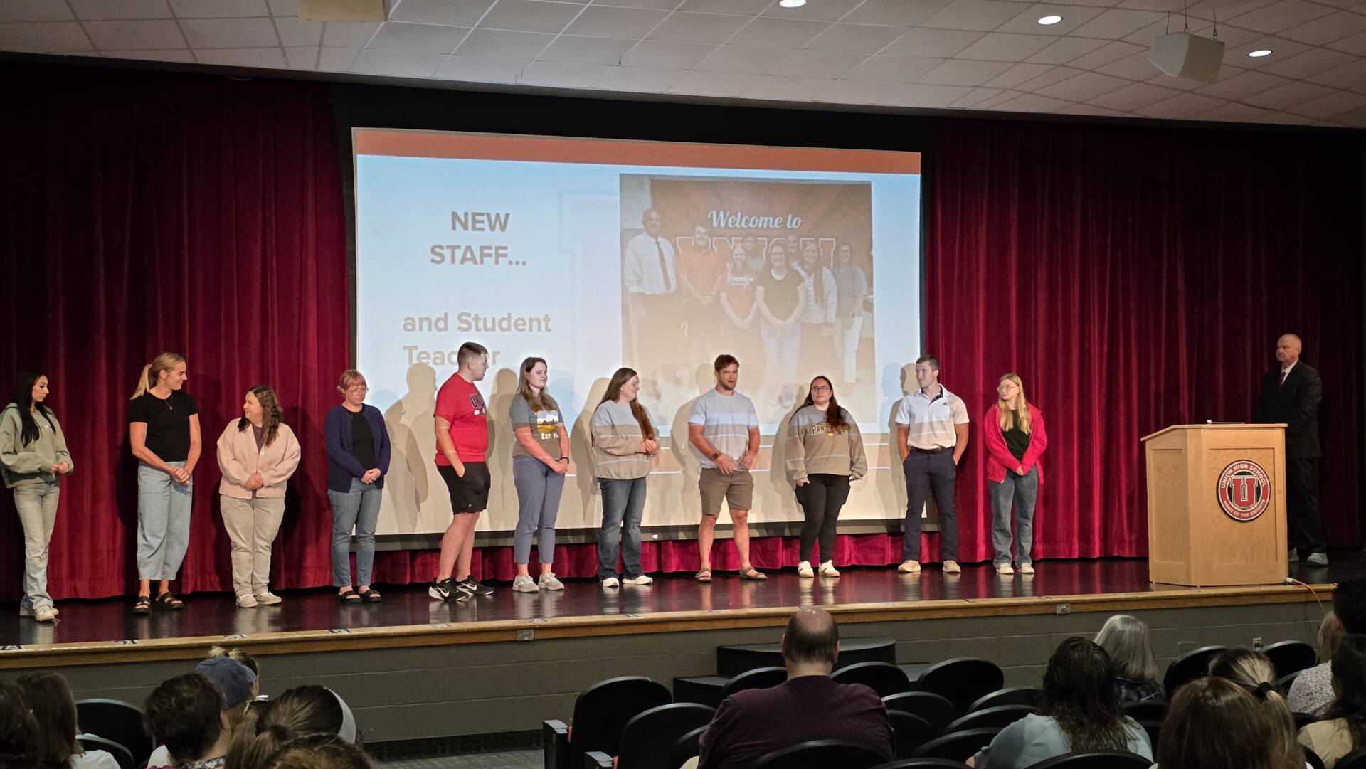 New staff members stand on an auditorium stage as they are introduced to a seated audience.
