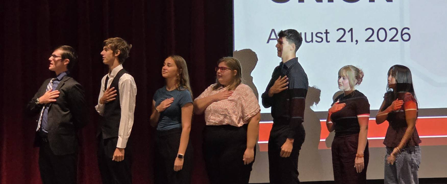 Seven students stand onstage with hands over their hearts during a school assembly on August 21, 2026.
