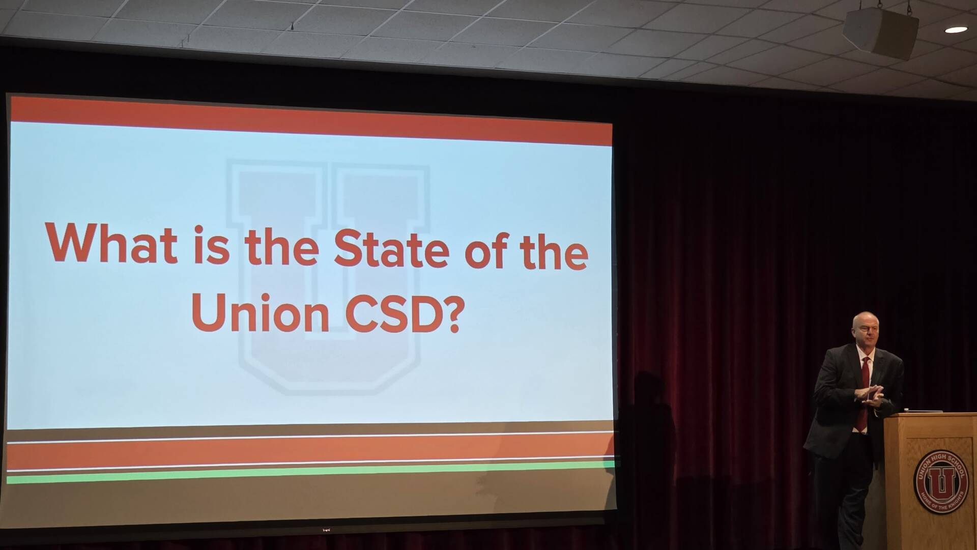 A presenter stands beside a projected slide reading, “What is the State of the Union CSD?” in a school auditorium.