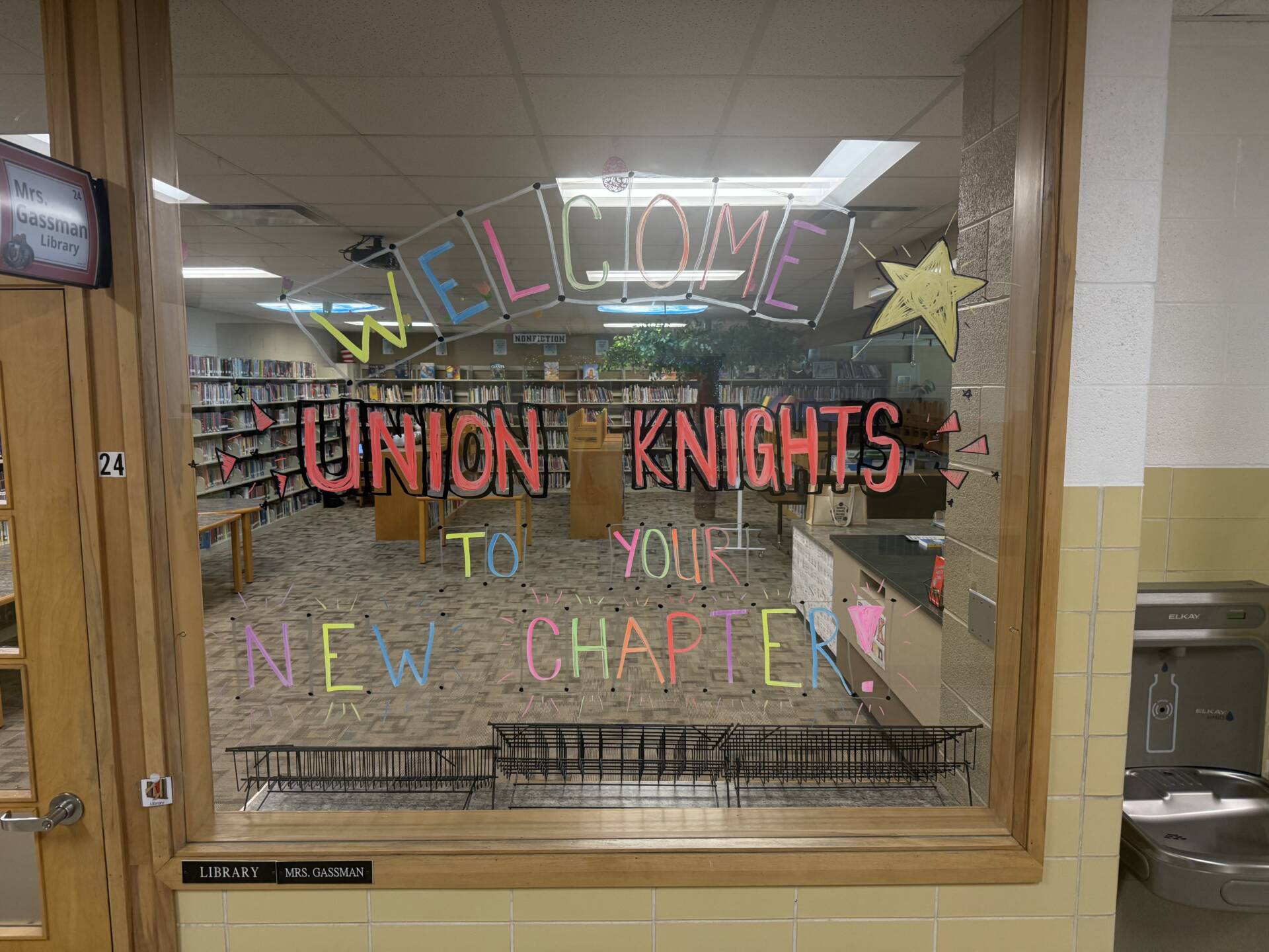 Colorful writing on a library window welcomes Union Knights “to your new chapter,” with bookshelves visible behind it.