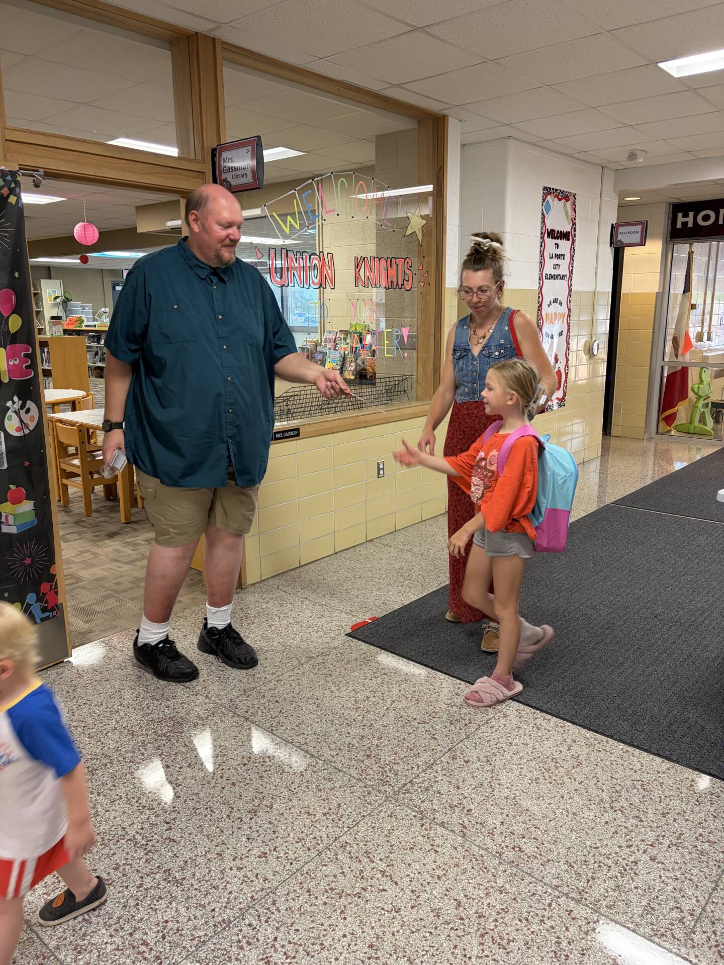 A student with a backpack reaches toward a staff member while an adult accompanies her through a welcoming school hallway.