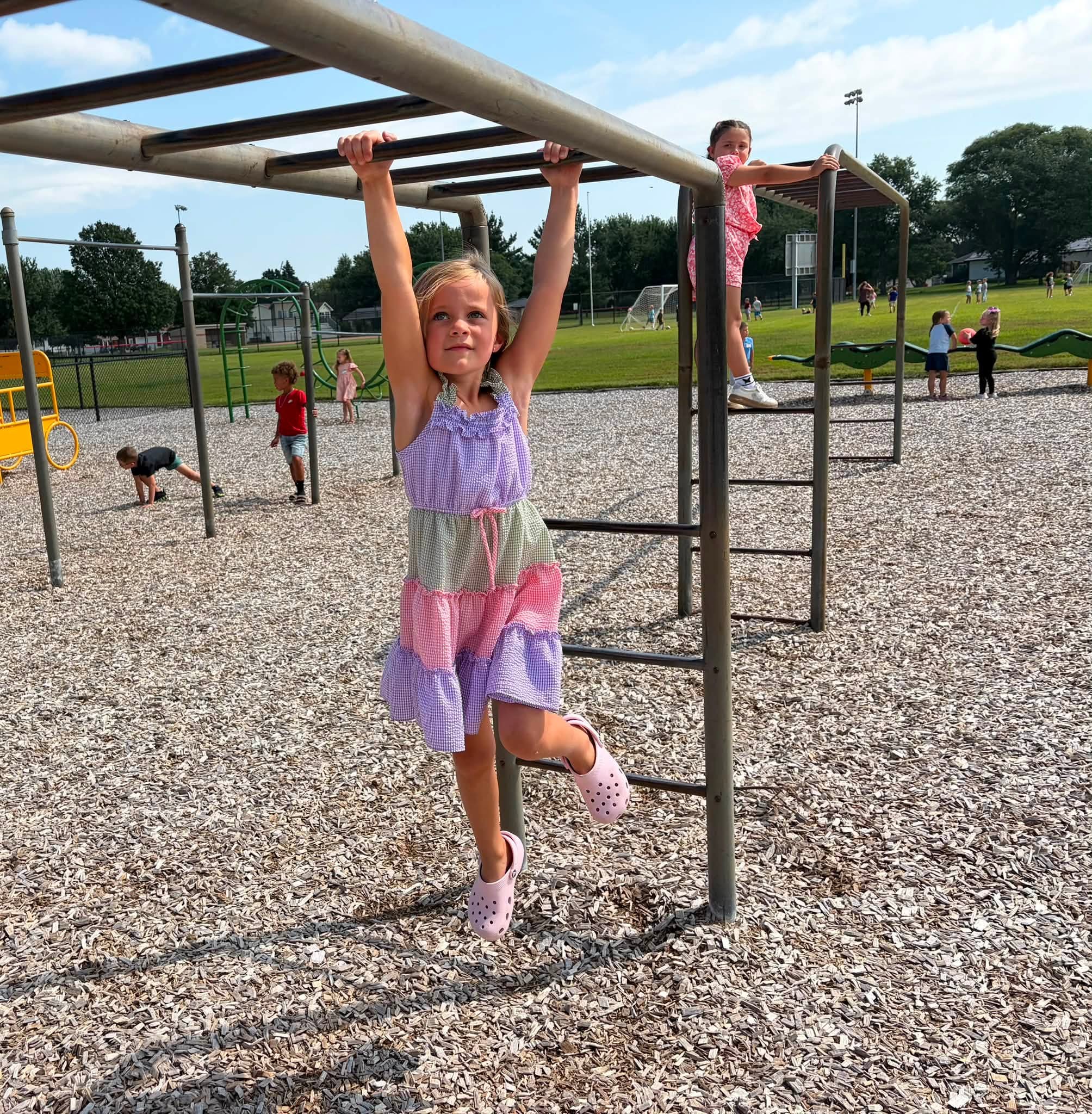 Student in a colorful dress hangs from monkey bars while classmates play across the sunny school playground.