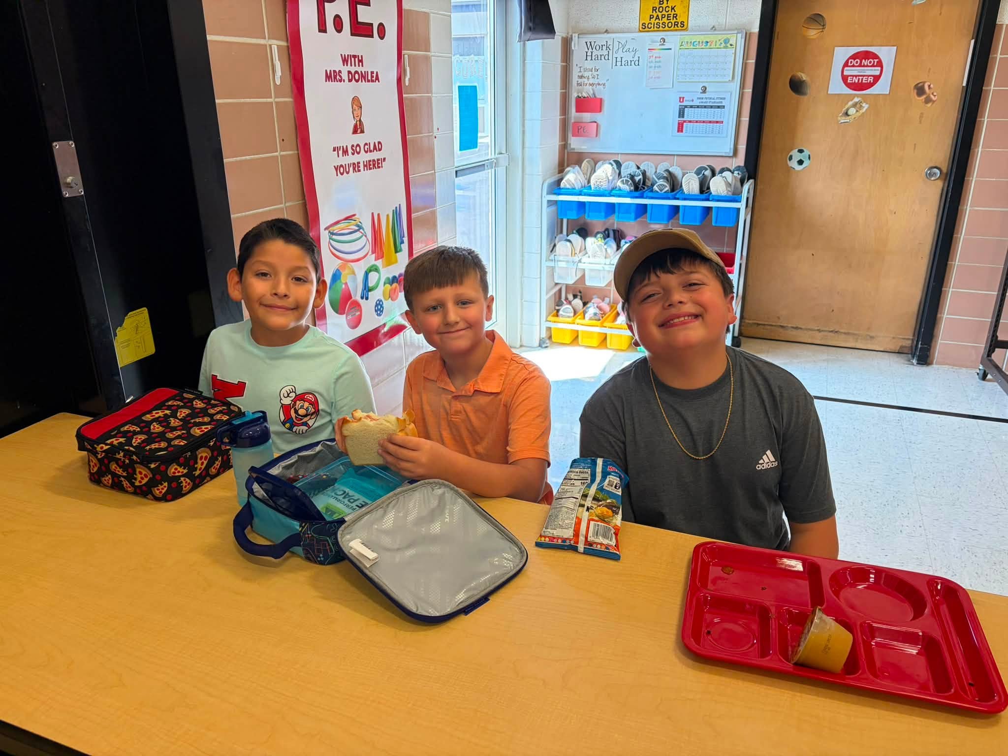 Three students smile at a cafeteria table with lunch boxes, food, and a red lunch tray.