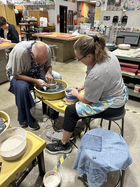 Two adults work side by side at pottery wheels, shaping clay in a classroom filled with ceramics supplies and equipment.