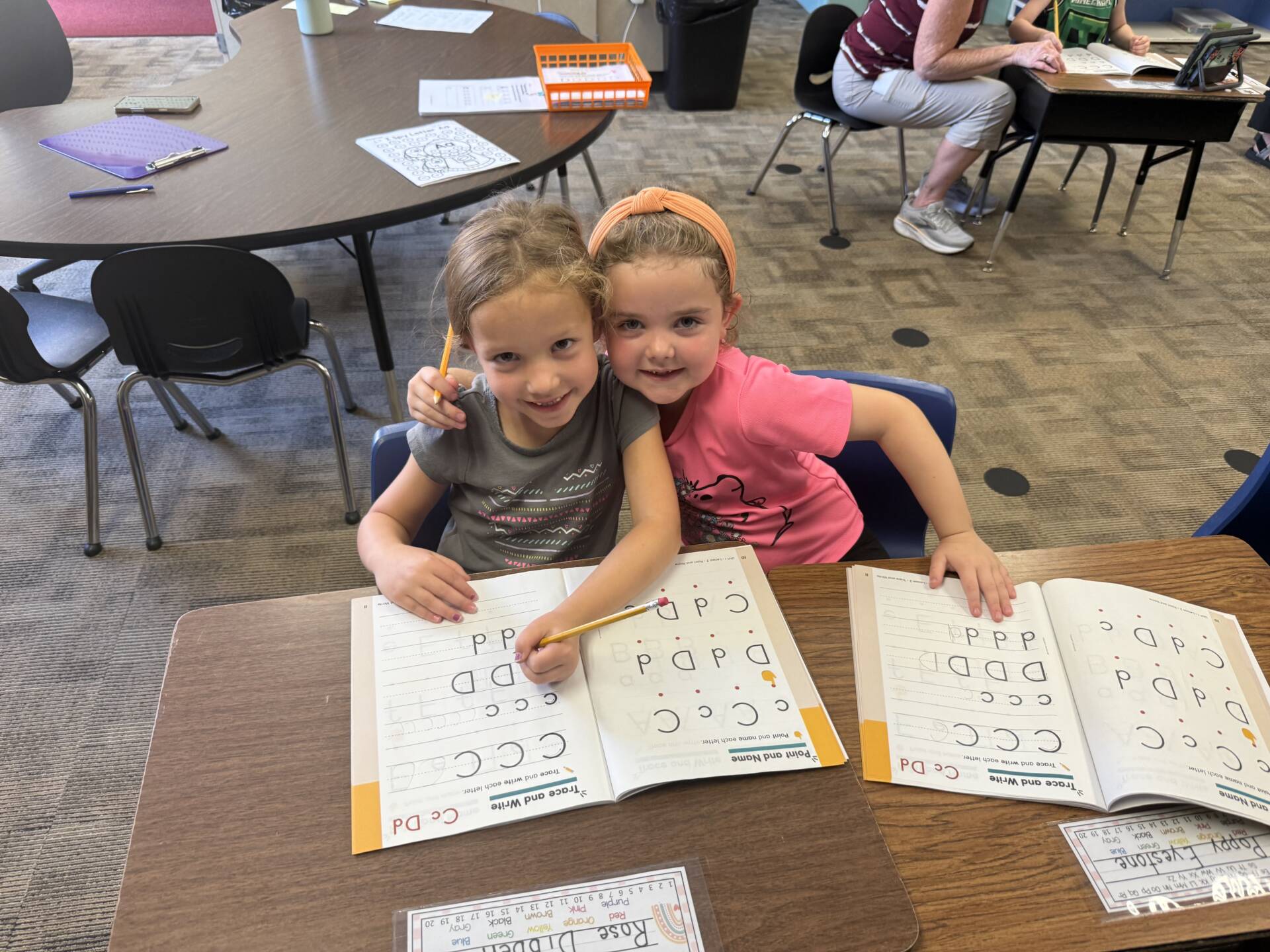 Two students sit closely together and smile while practicing letter formation in handwriting workbooks.