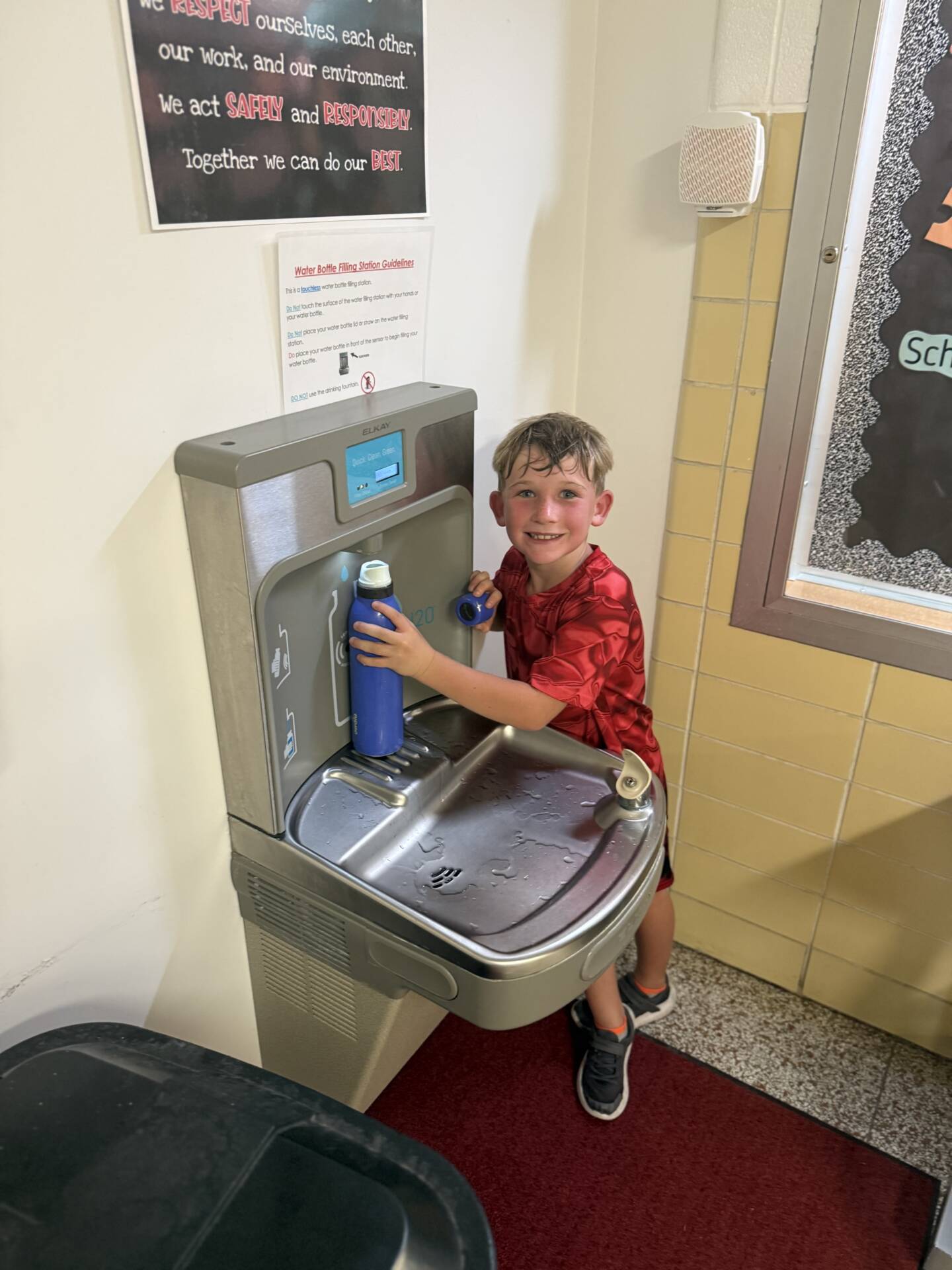 Smiling student fills a blue water bottle at a school drinking fountain and bottle-filling station.
