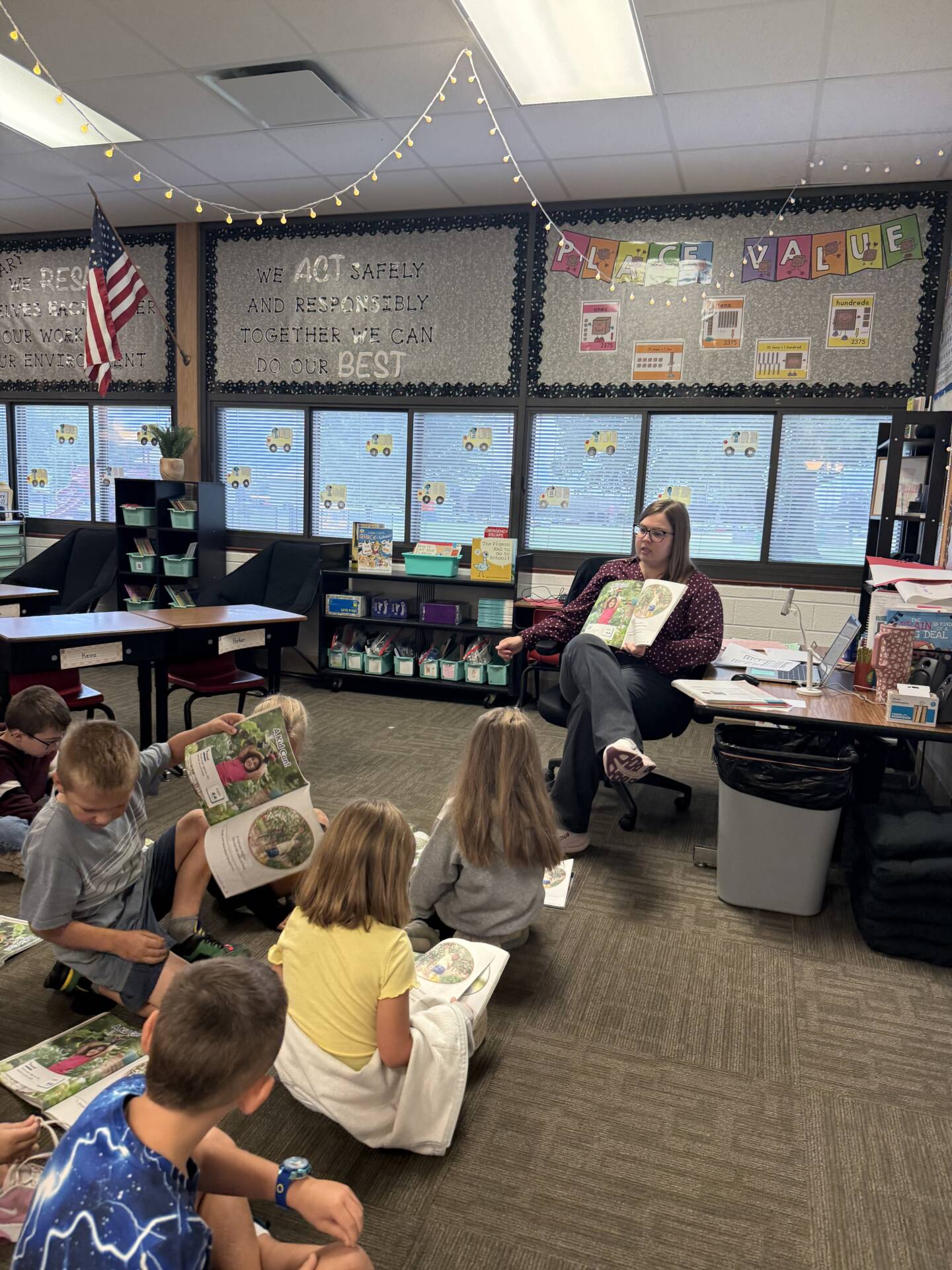 A teacher reads aloud while students sit on the classroom floor and follow along in their own books.