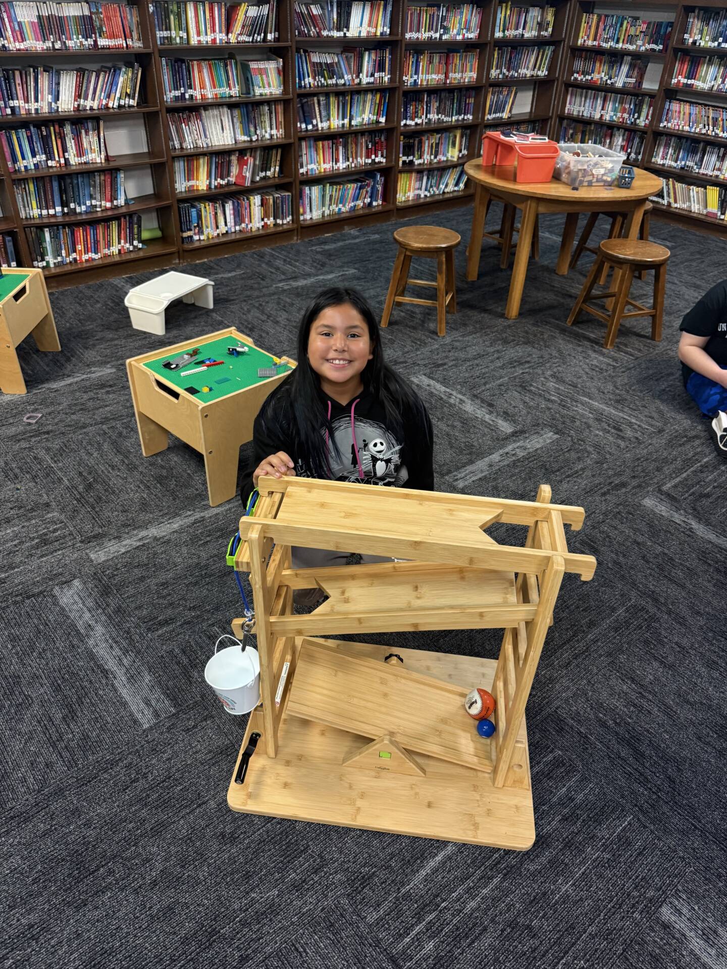 A smiling student poses behind a wooden activity structure in a school library surrounded by bookshelves.