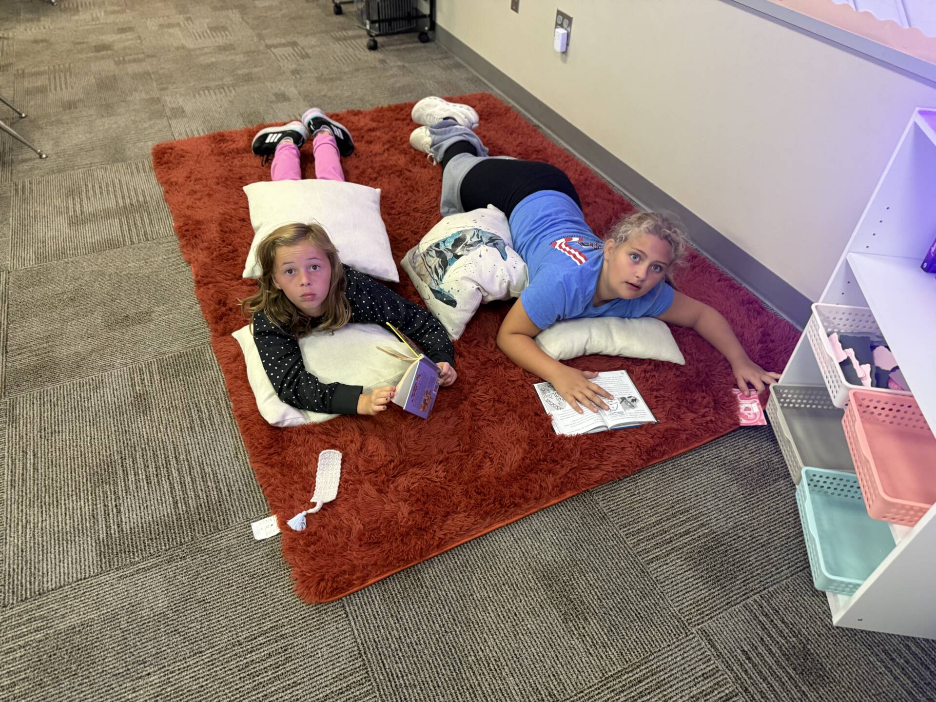Two students lie on pillows on a red classroom rug while reading books together.