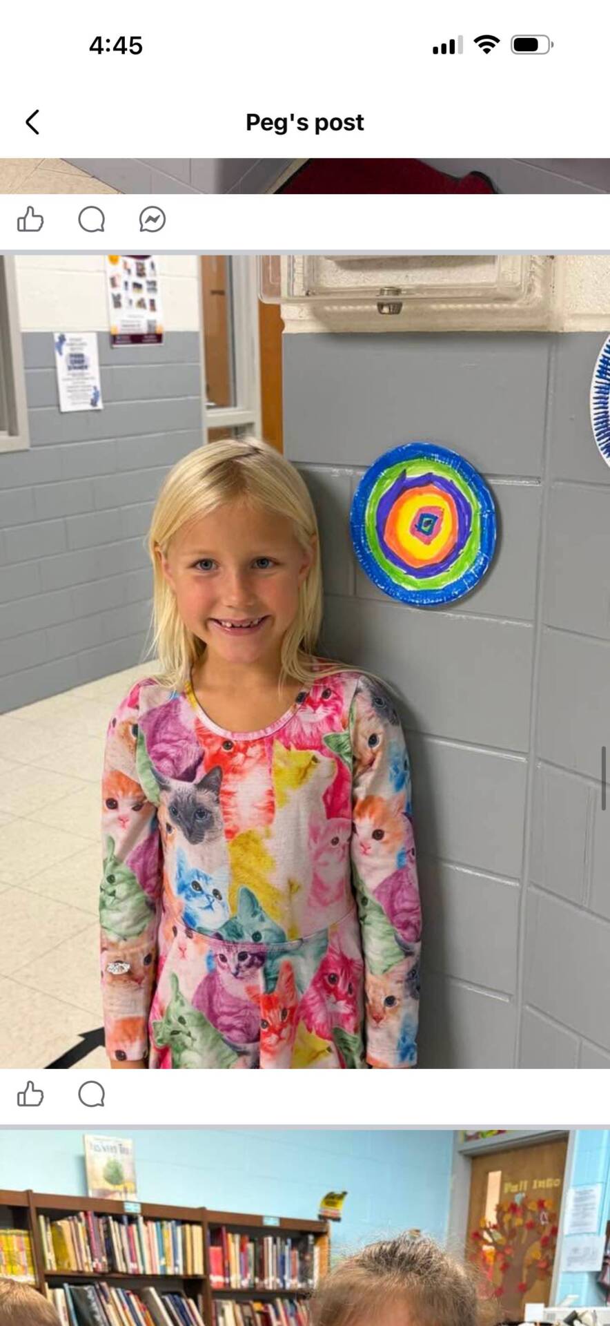 Smiling elementary student stands beside a colorful concentric-circle artwork displayed on a school hallway wall.