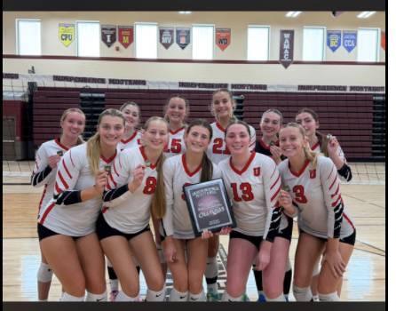 A girls’ volleyball team poses together in a gym while holding a tournament champions plaque.