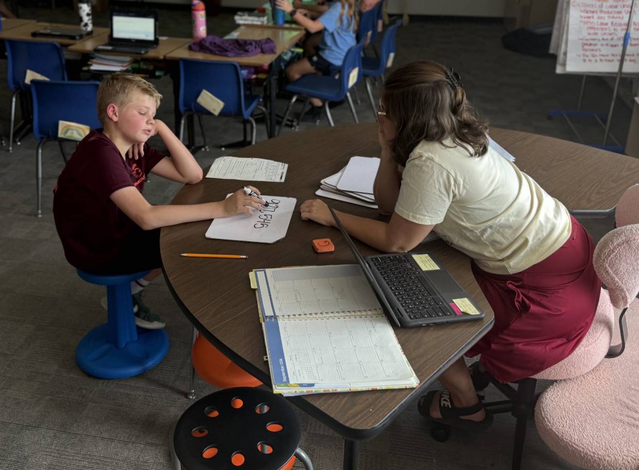 A student writes on a small whiteboard while working one-on-one with a teacher at a classroom table.