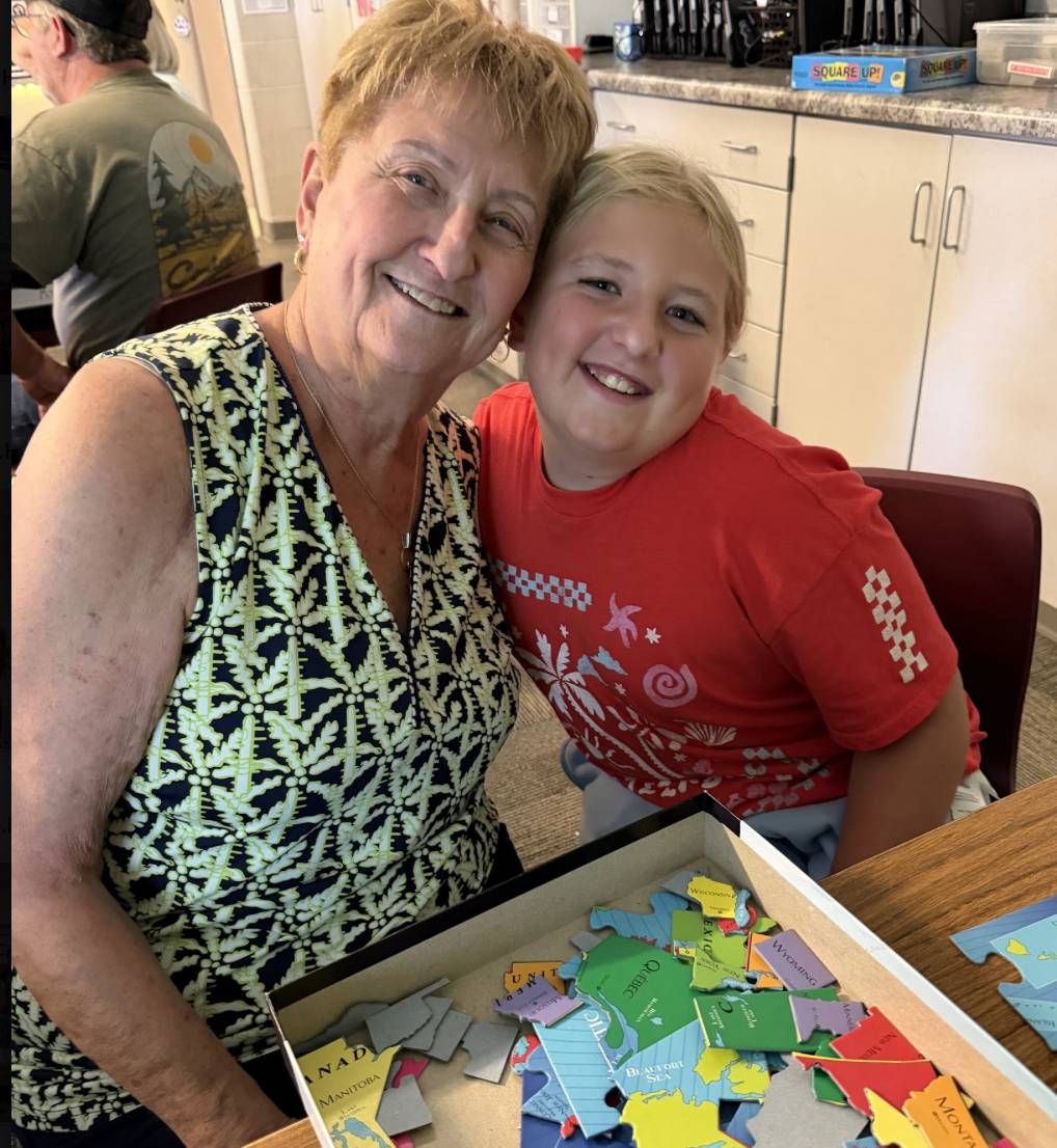 A smiling student and older adult lean together beside a tray filled with colorful map puzzle pieces.