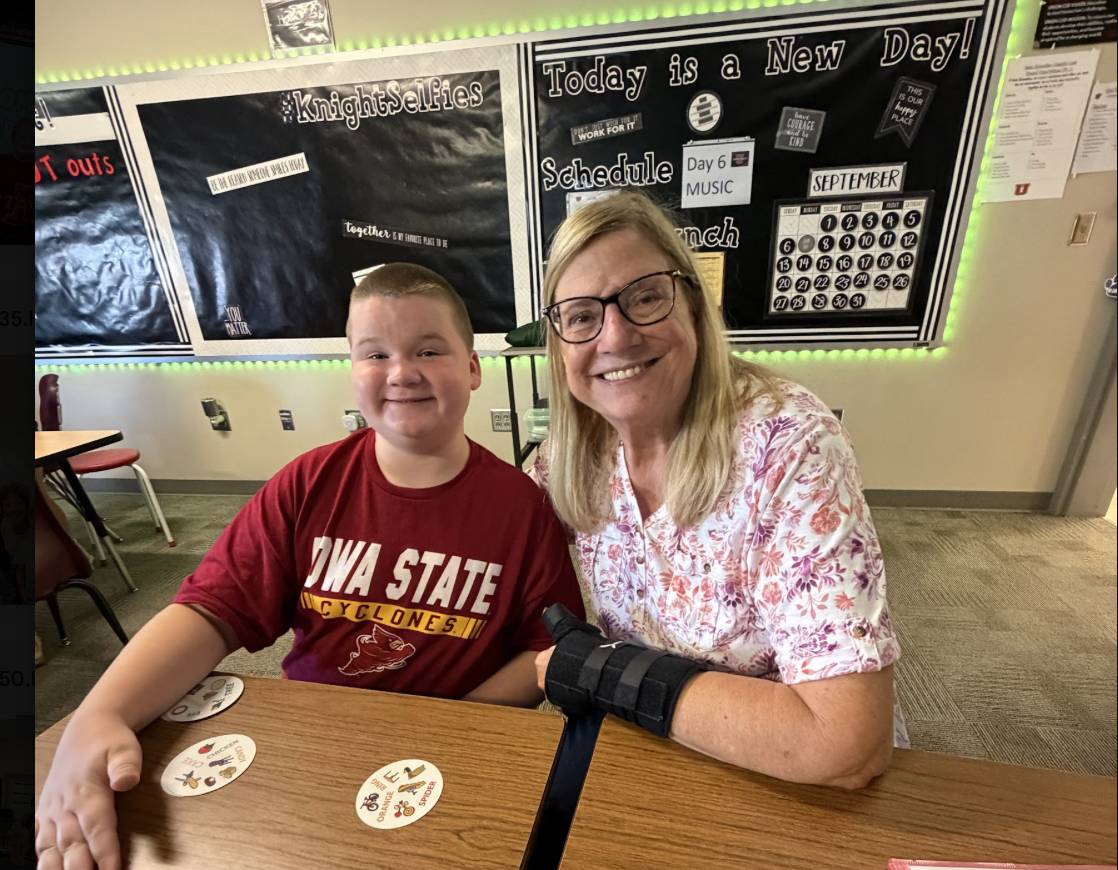 A student and an older adult smile together at a classroom desk with learning materials nearby.