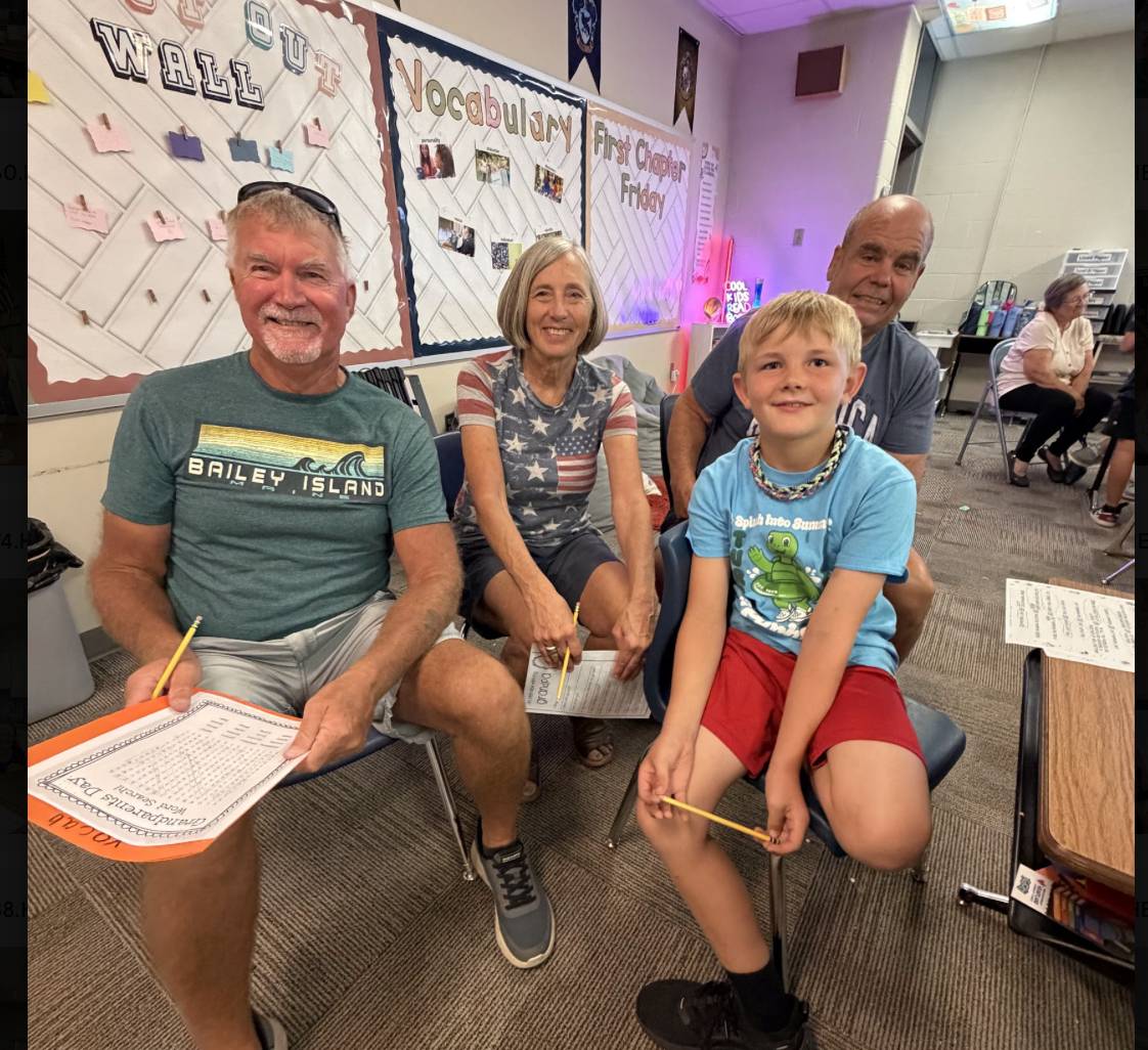 A student poses with three older adults holding pencils and activity sheets in a classroom.