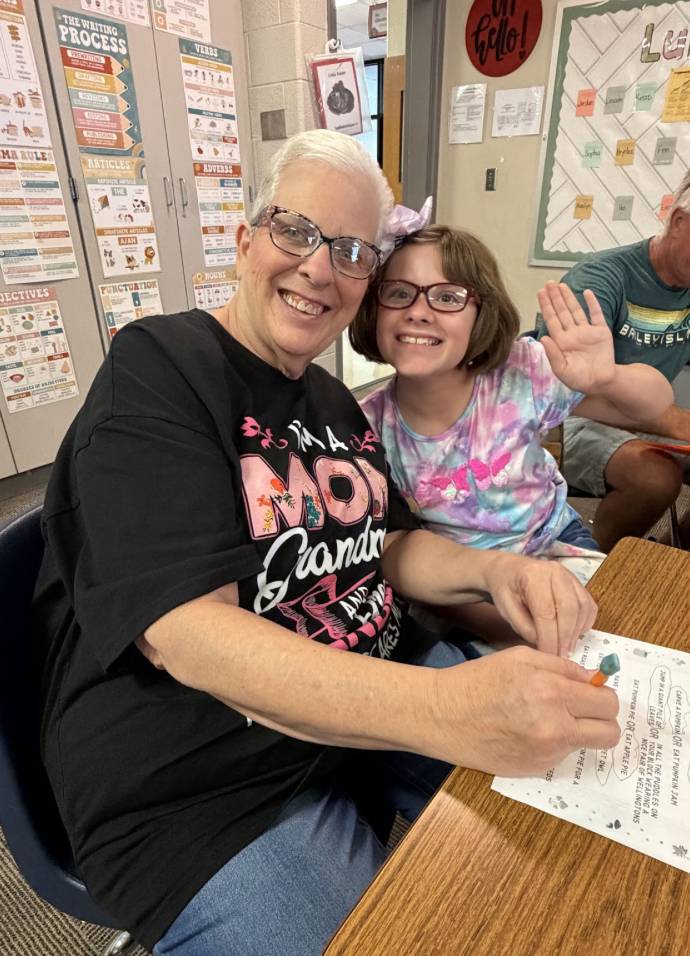A smiling older adult and student embrace at a classroom desk as the student waves toward the camera.