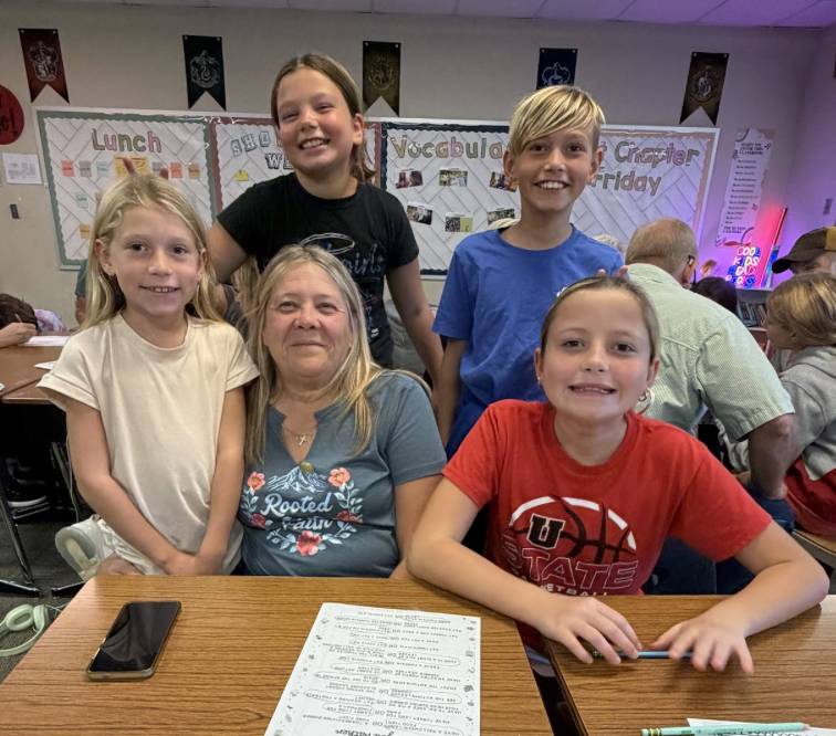Four students gather around a seated older adult and smile for a group photo at a classroom table.