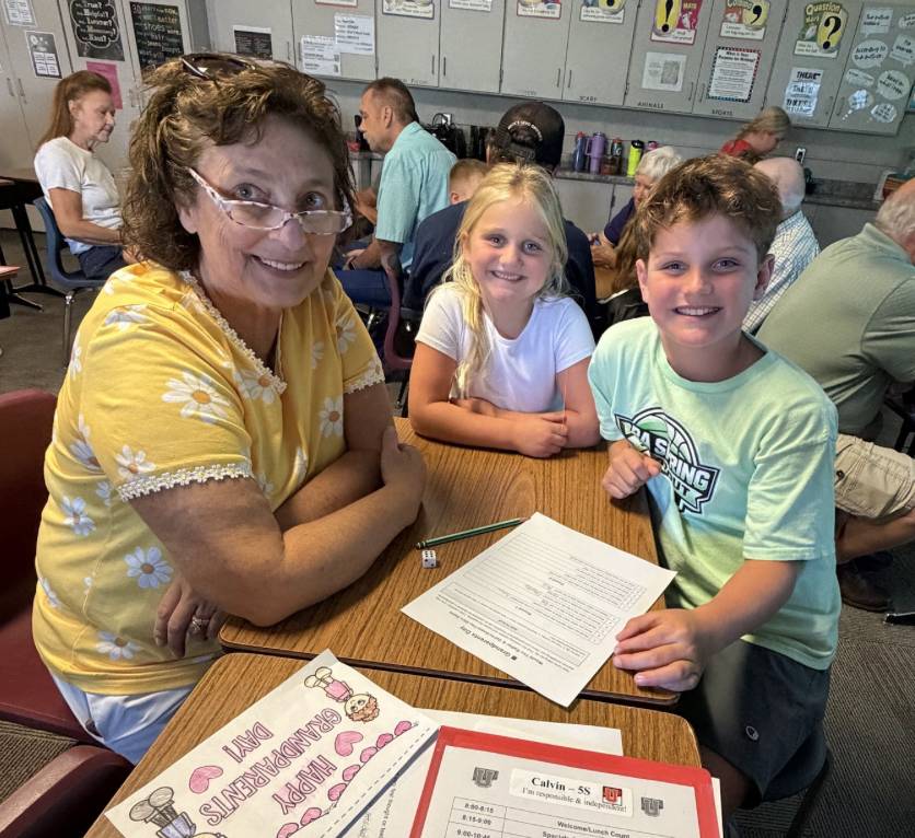 An older adult and two students smile together at a classroom table with worksheets and pencils.