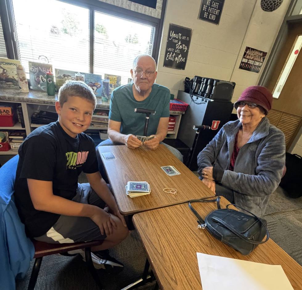 A student and two older adults sit around a classroom table with cards laid out for a game.