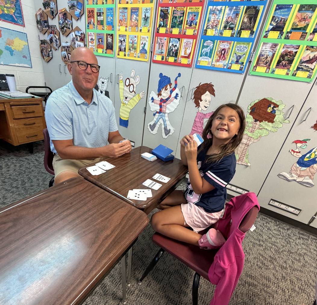 A student and an older adult smile while playing a card game together at a classroom table.