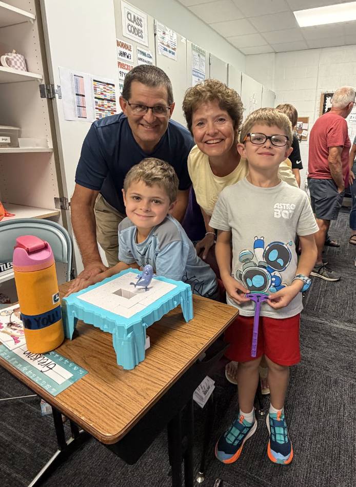 Two older adults and two smiling students pose together beside a classroom table with a learning activity.