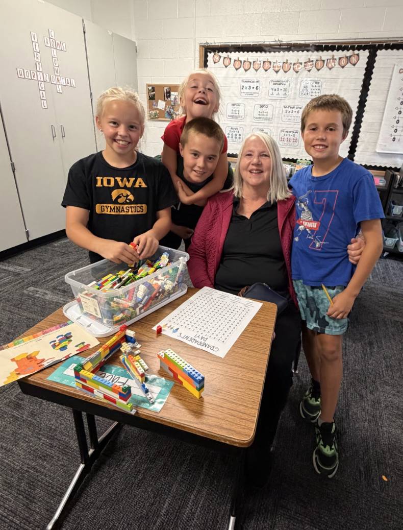Four smiling students gather around an older adult seated at a classroom table covered with colorful building blocks.