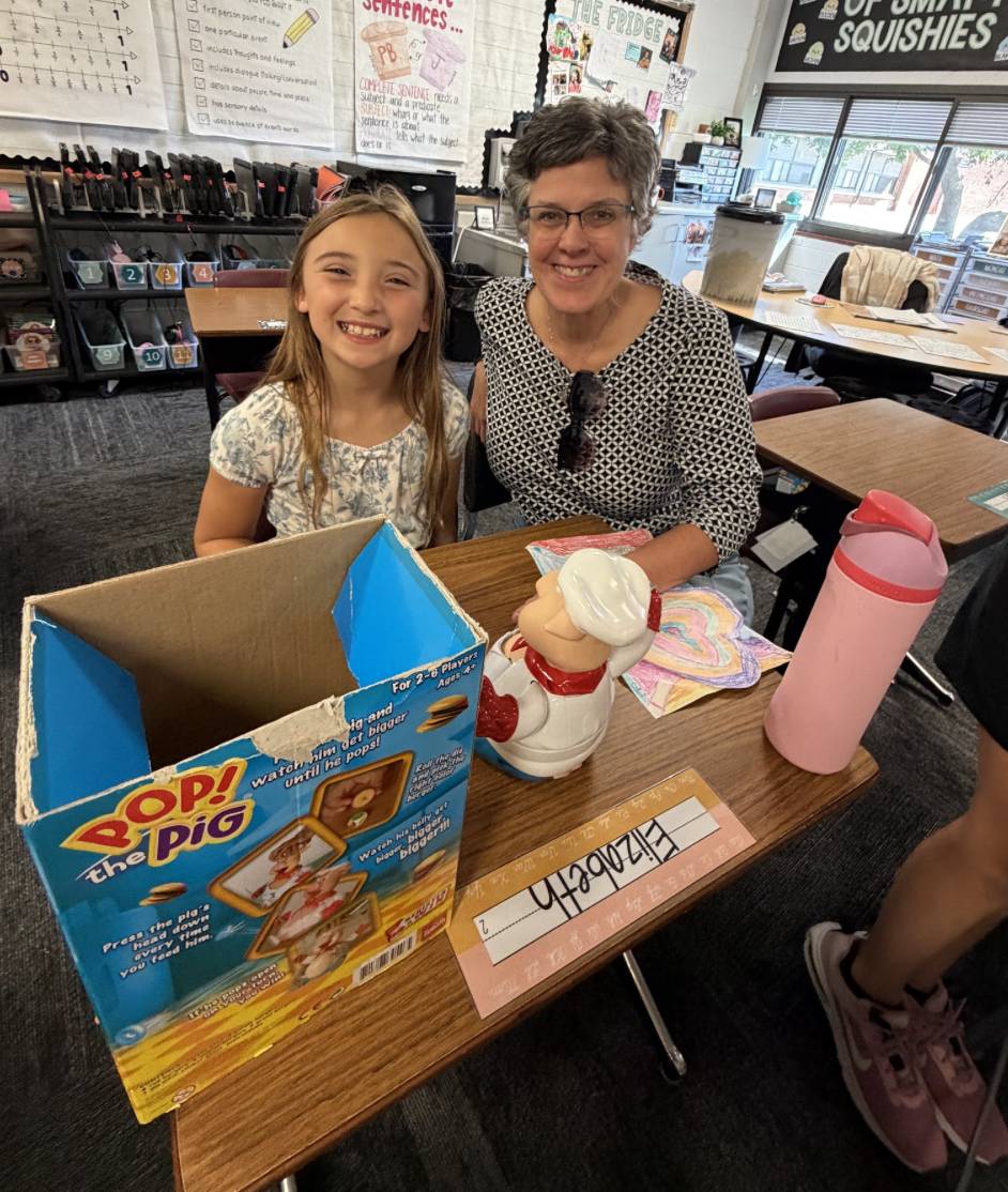 A smiling student and older adult sit together at a classroom desk with a game, artwork, and water bottle.