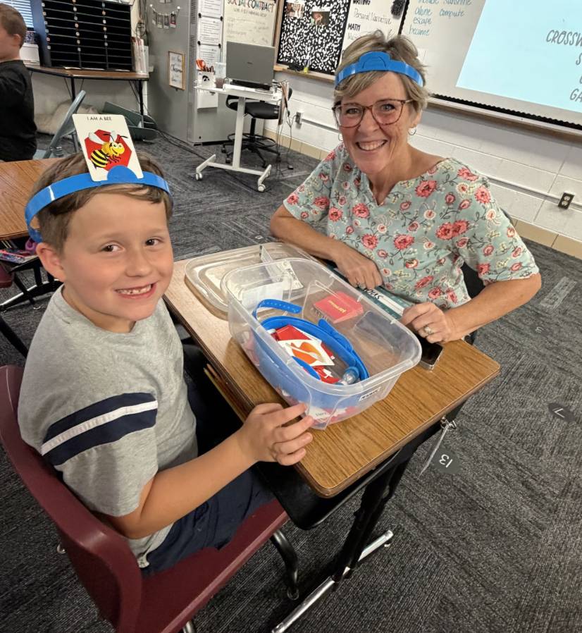 A smiling student and older adult wear blue headbands while playing a classroom guessing game together.