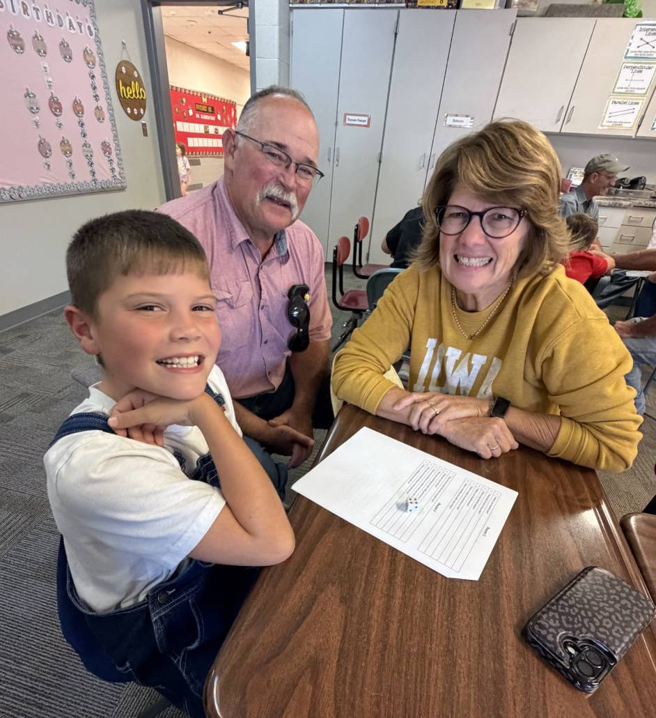 A smiling student poses with two older adults at a classroom table with a worksheet and die.