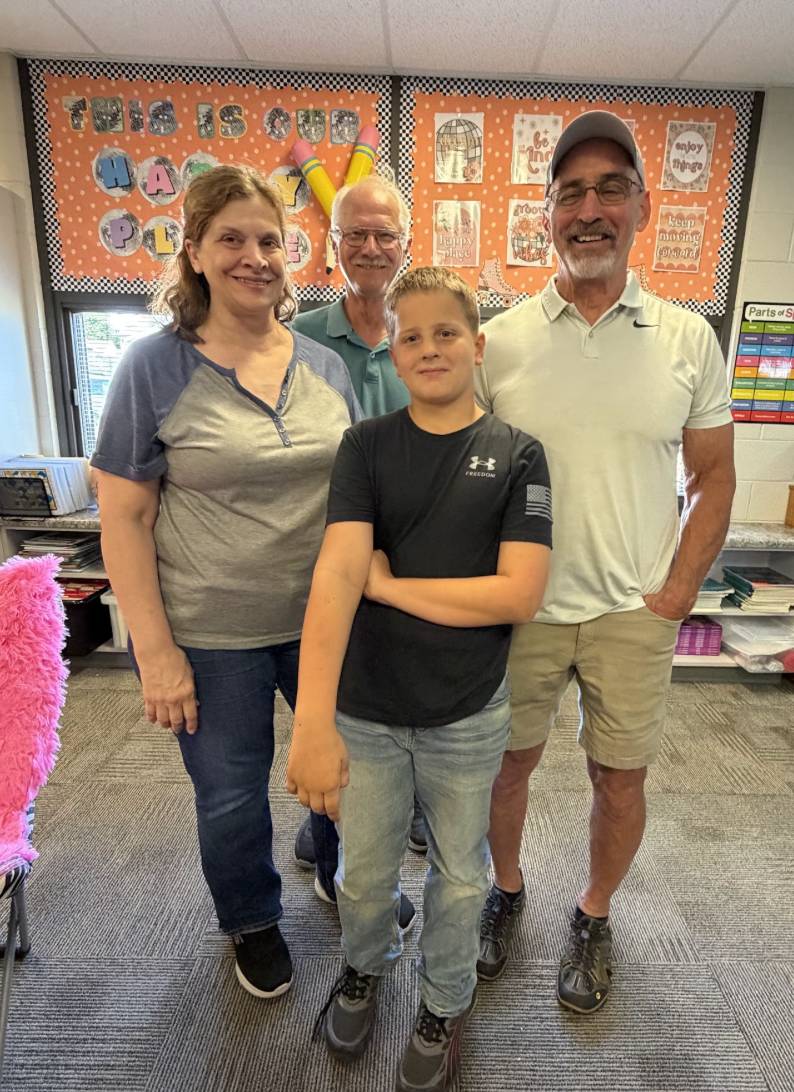 A student poses with three older adults in front of a colorful classroom bulletin board.