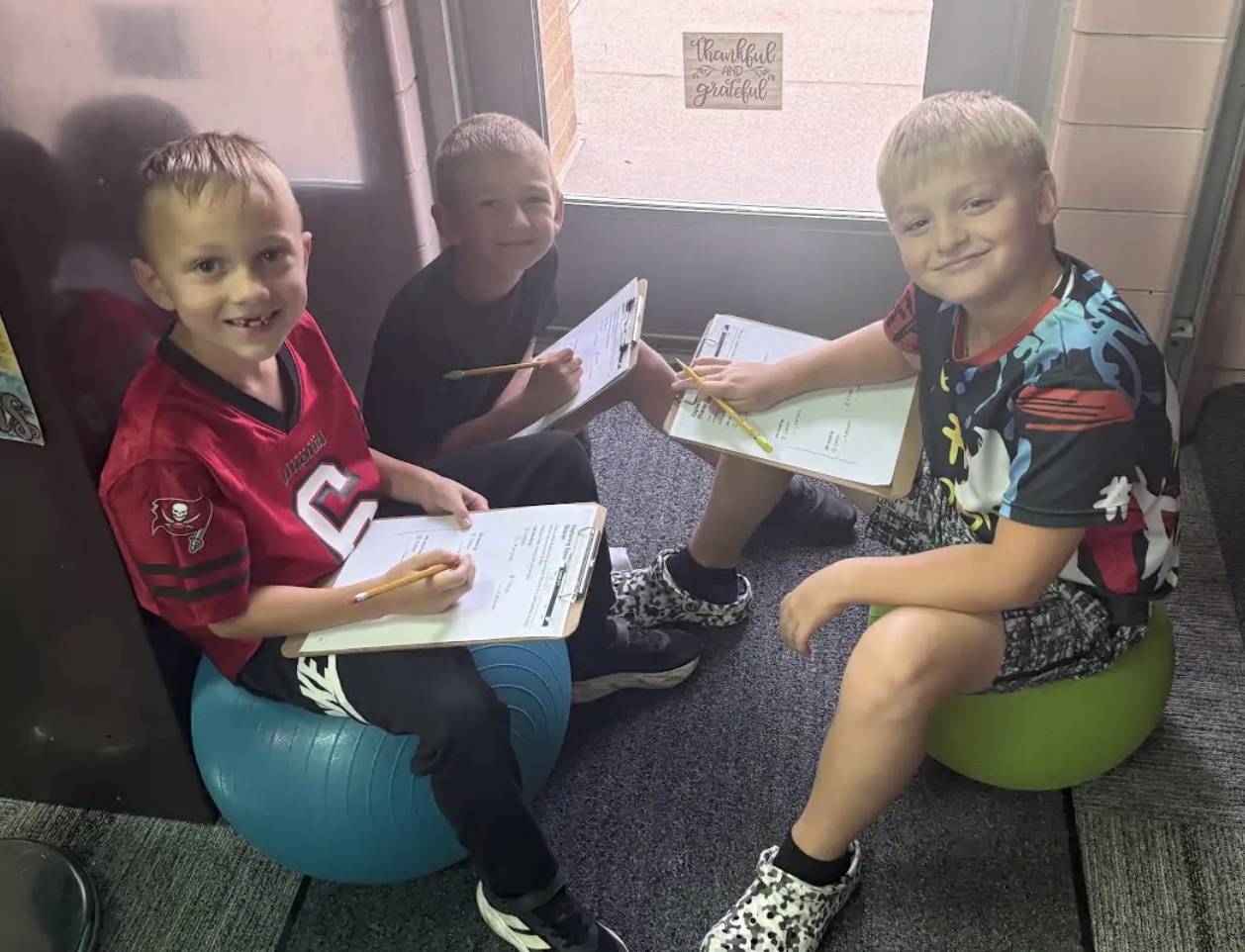 Three students sit on flexible classroom seats with clipboards and pencils while working together.