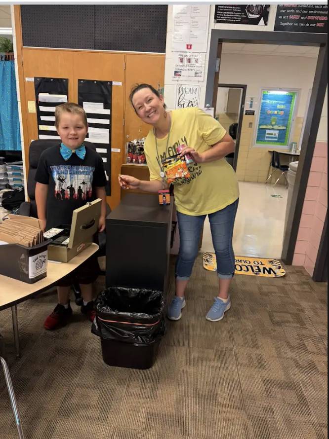 A smiling student and adult stand together in a classroom holding small treats during a school activity.