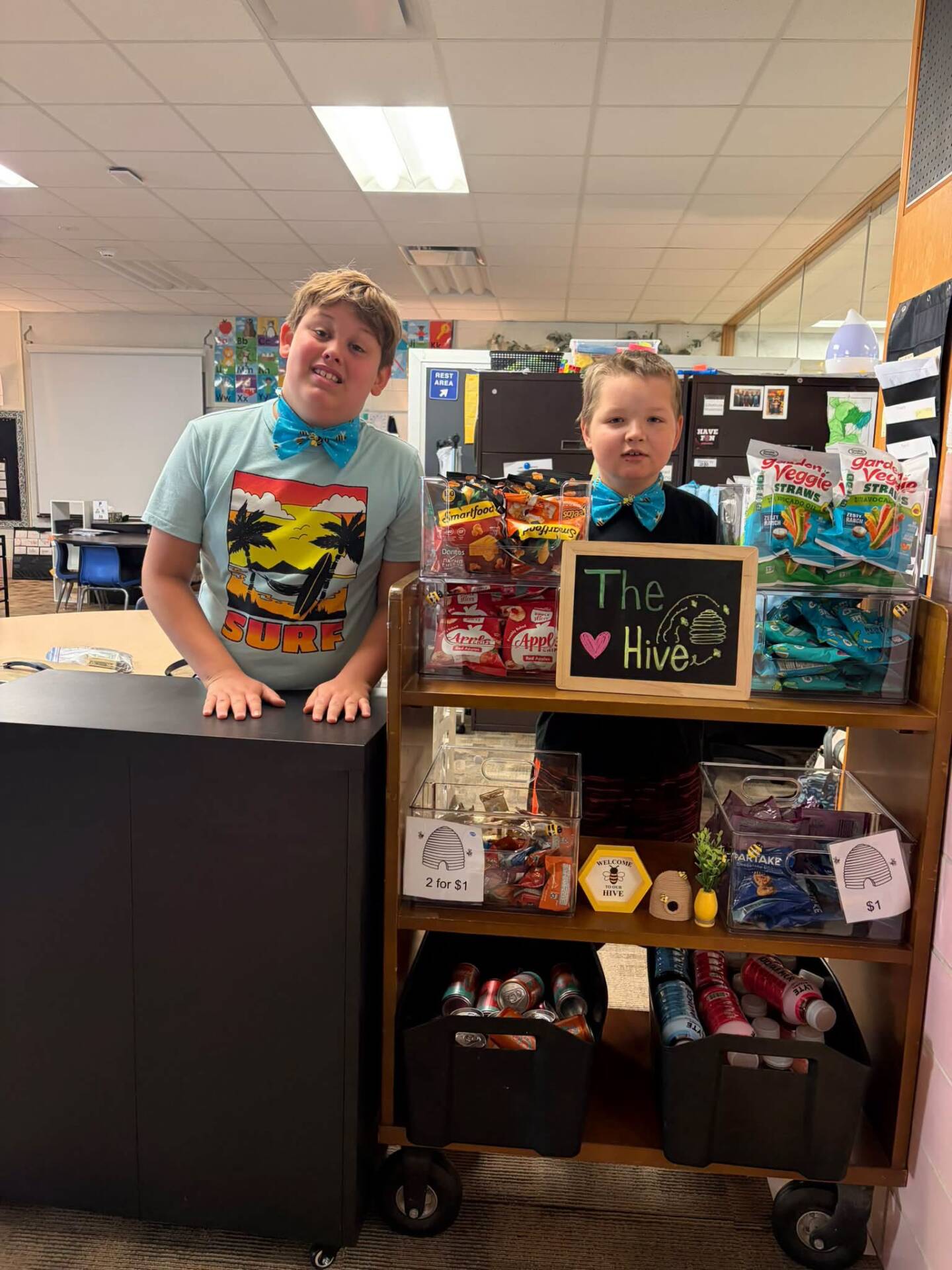 Two boys wearing colorful bow ties pose behind a school snack cart labeled “The Hive.”