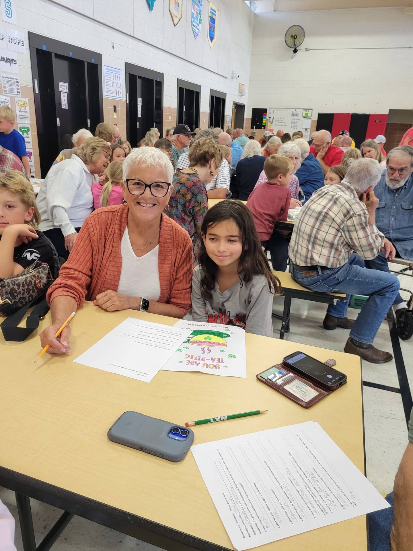 An older woman and a child sit together at a table during a busy school gathering, smiling beside activity sheets.
