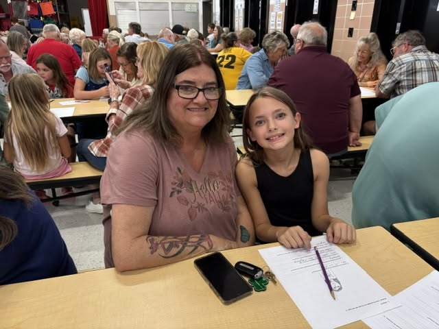 An adult and a child smile together at a crowded school gathering, seated at a table with activity papers.
