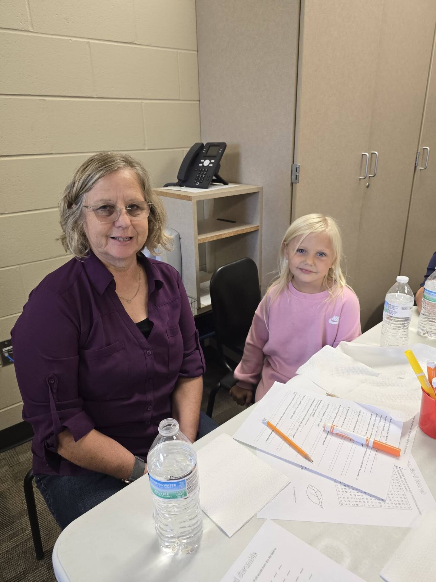 An older woman and a young child smile together at a table covered with school activity sheets and supplies.
