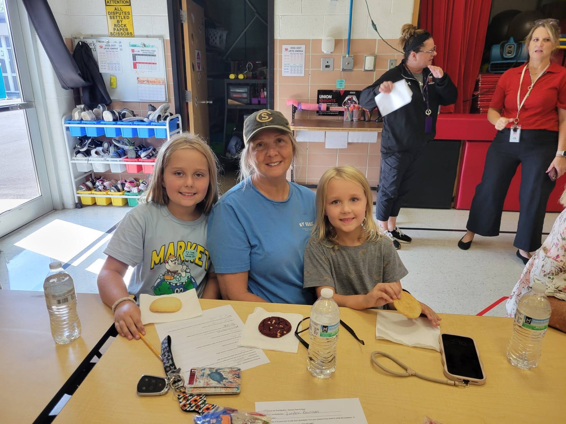 An adult and two children smile at a table with snacks, water bottles, and school activity sheets.