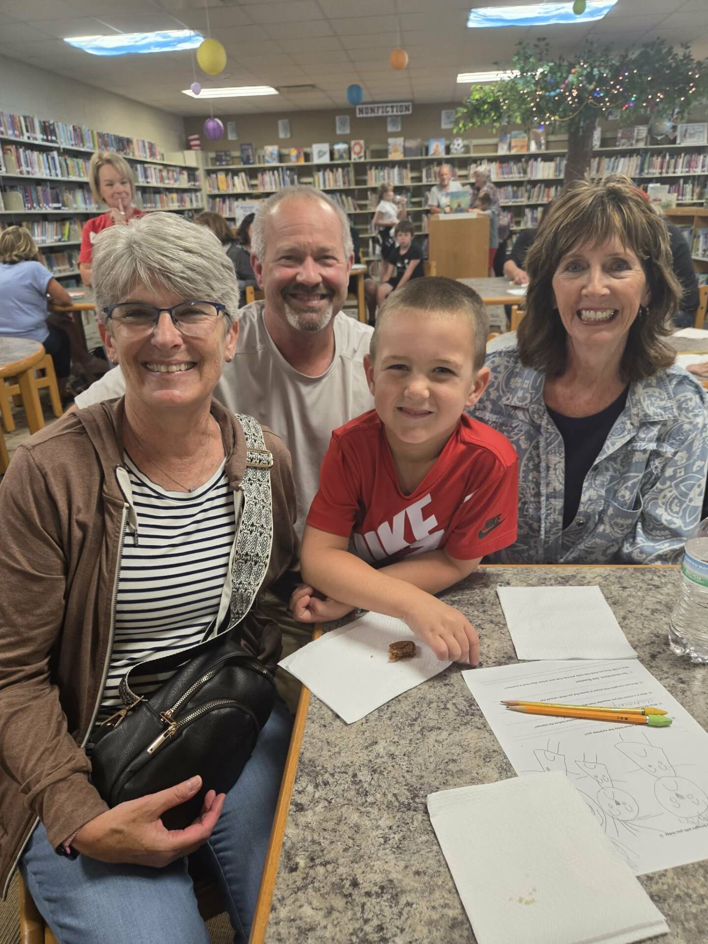 A child in a red shirt smiles with three adults around a table during a lively school library gathering.