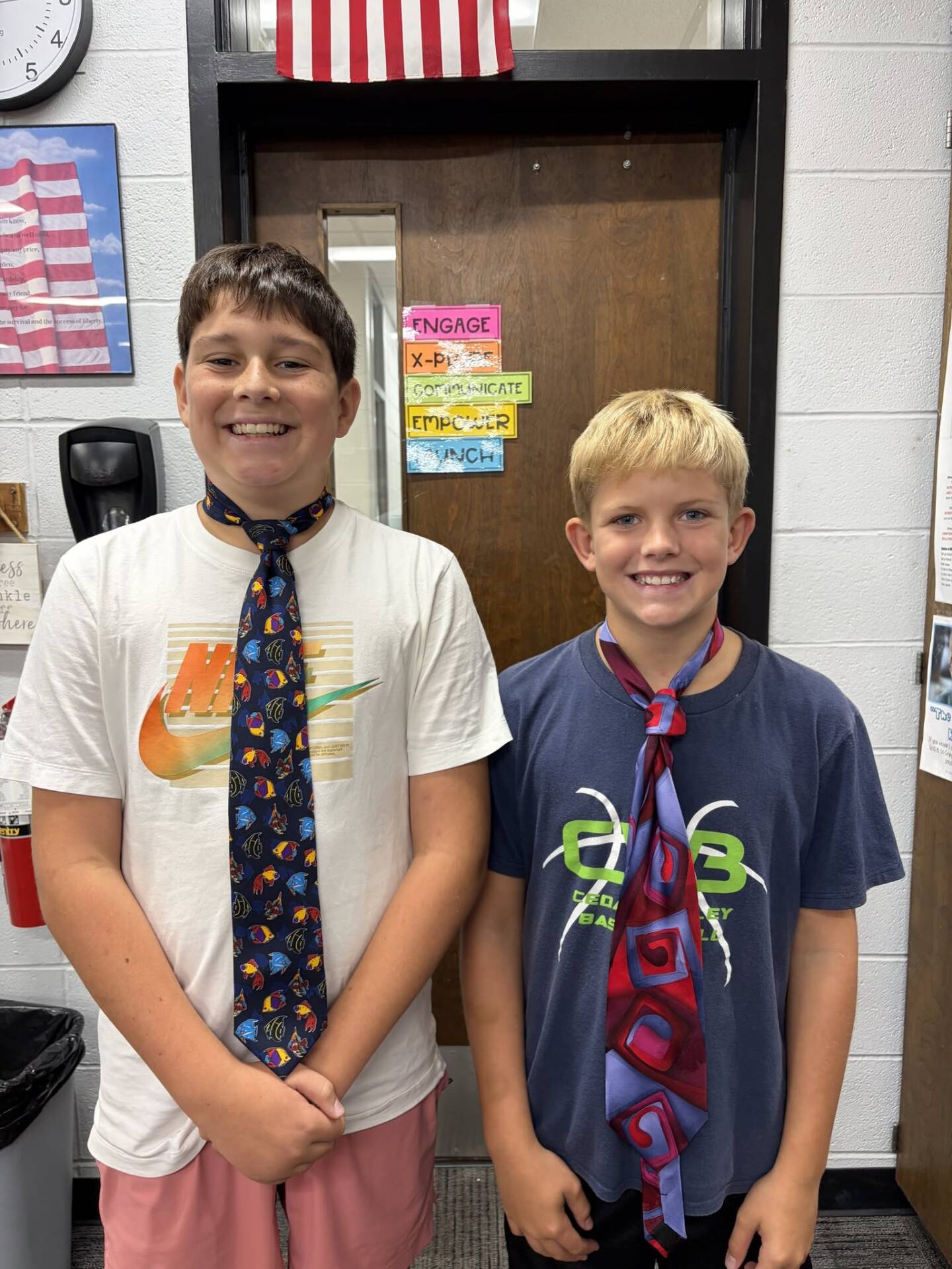 Two smiling boys wearing colorful neckties pose together in a school classroom.