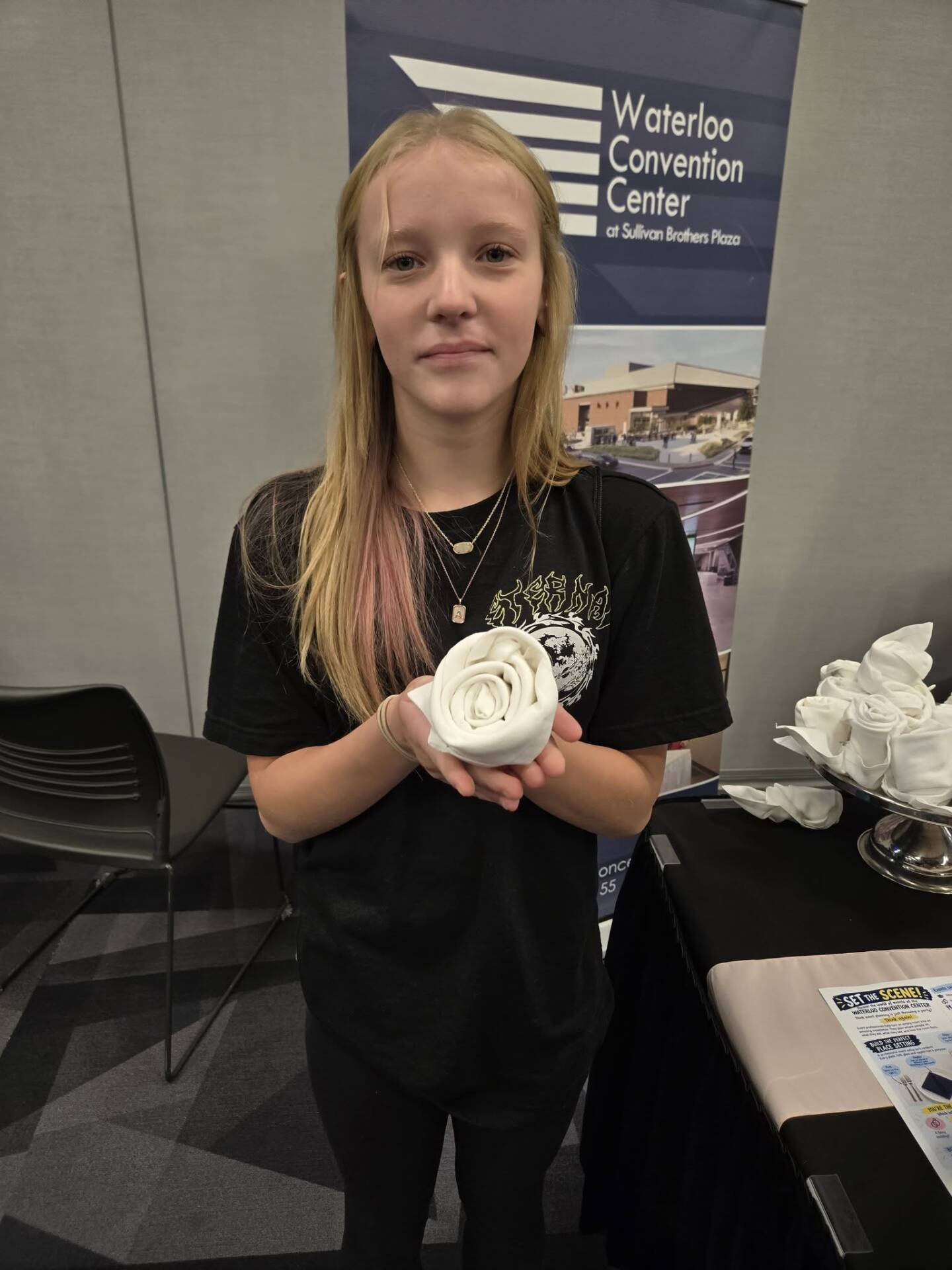 A girl holds a white fabric rose she created while standing at an event display.