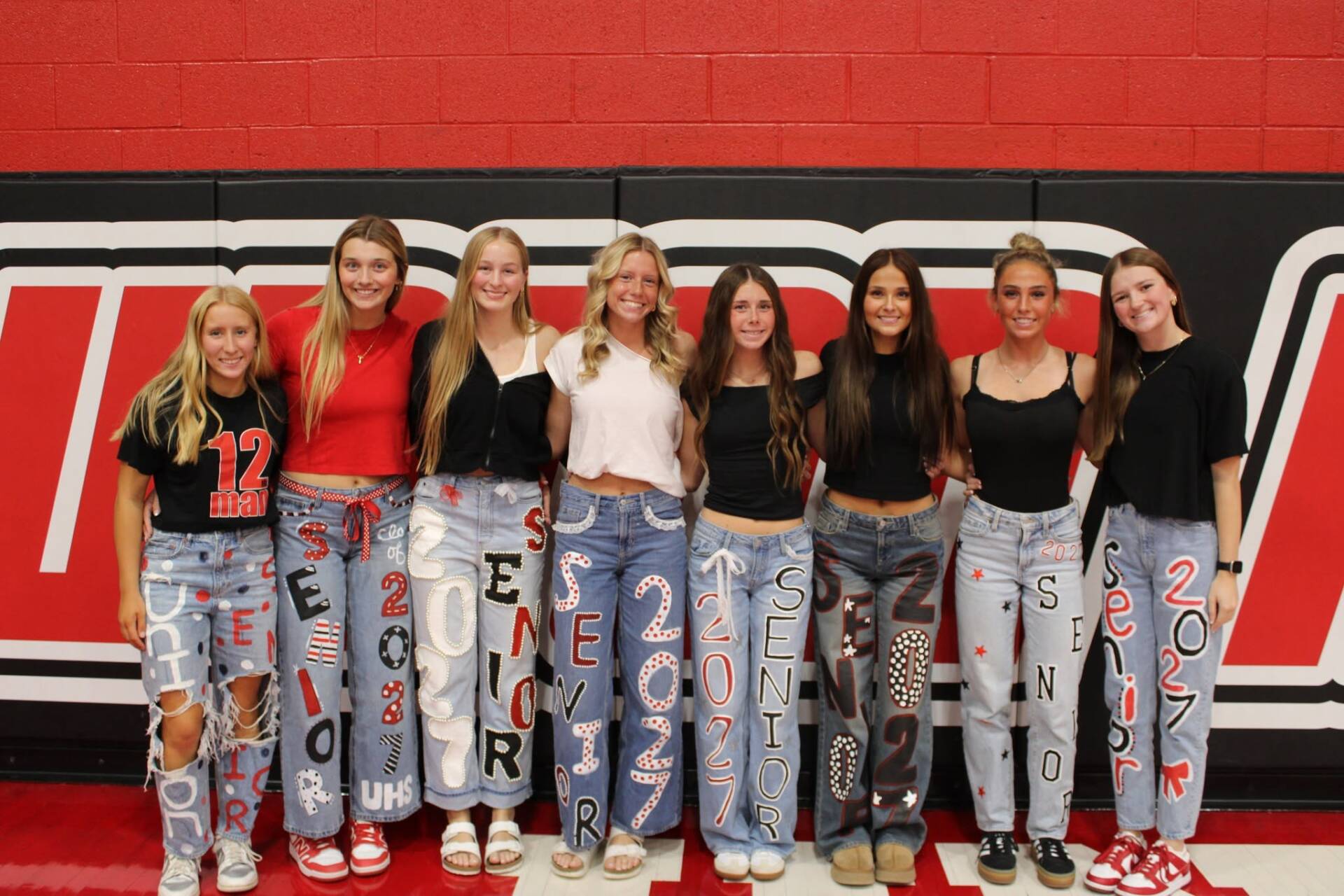 Eight high school seniors pose together in decorated jeans celebrating the Class of 2027 against a red and black gym wall.