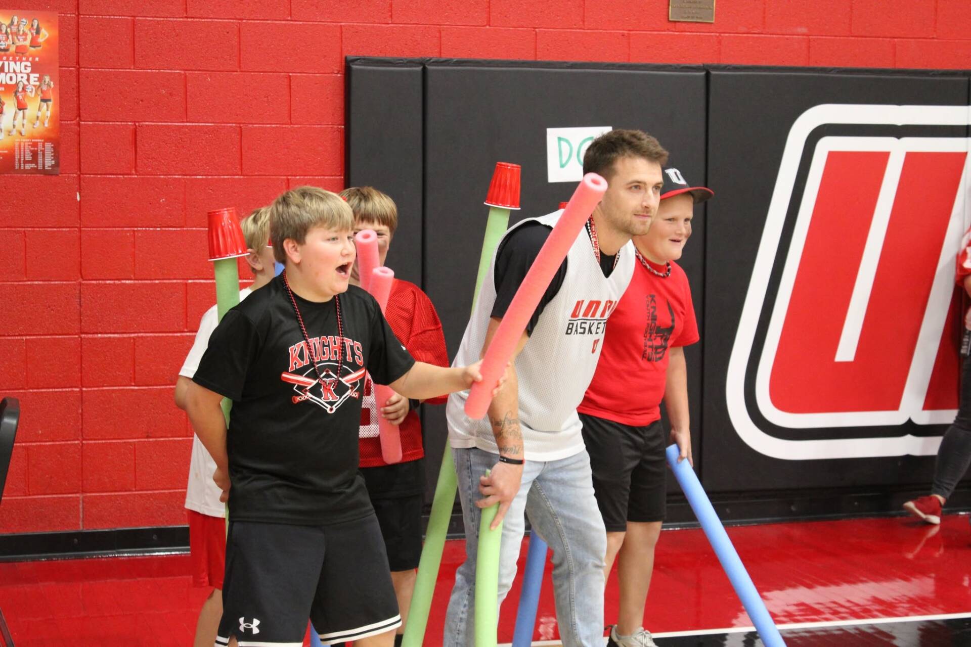 Students and an adult participate in a gym activity, holding colorful foam noodles and cheering together.
