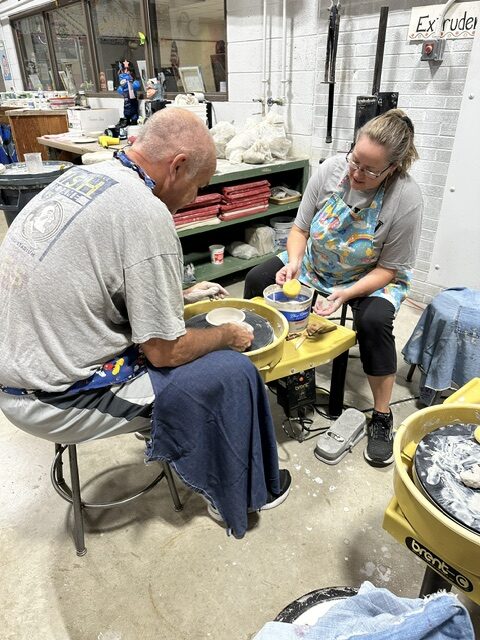 Two adults shape clay at pottery wheels while working together in a busy ceramics studio.