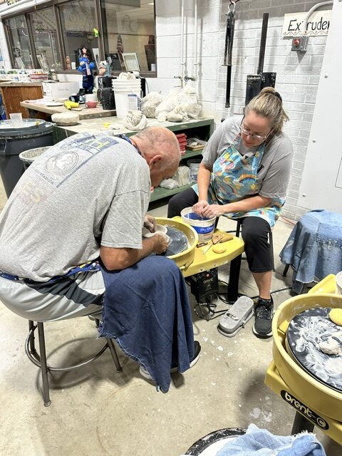 Two adults work together at pottery wheels in a ceramics studio filled with clay and art supplies.