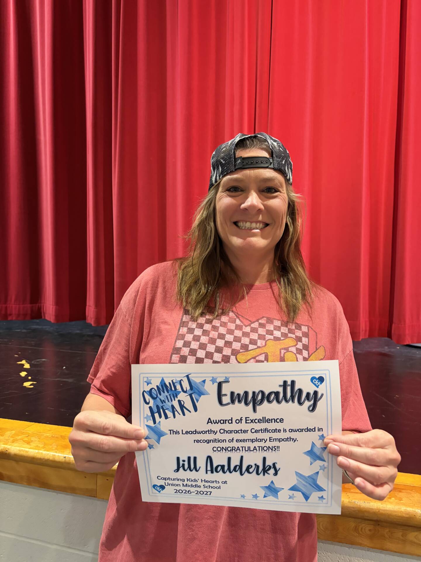 Jill Aalderks smiles while displaying her Empathy Award of Excellence certificate in front of a red stage curtain.