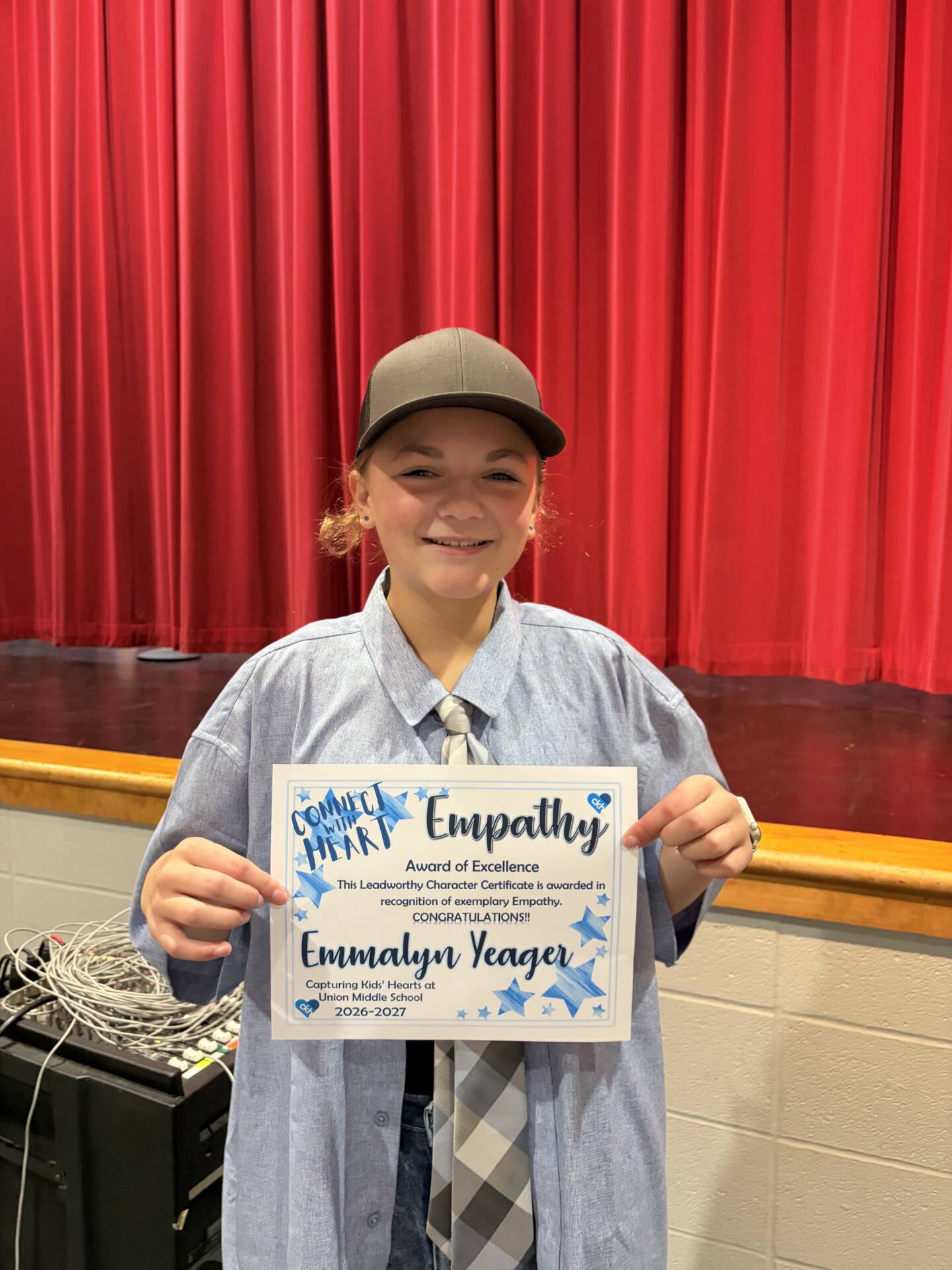 Emmalyn Yeager smiles while holding her Empathy Award of Excellence certificate in front of a red stage curtain.