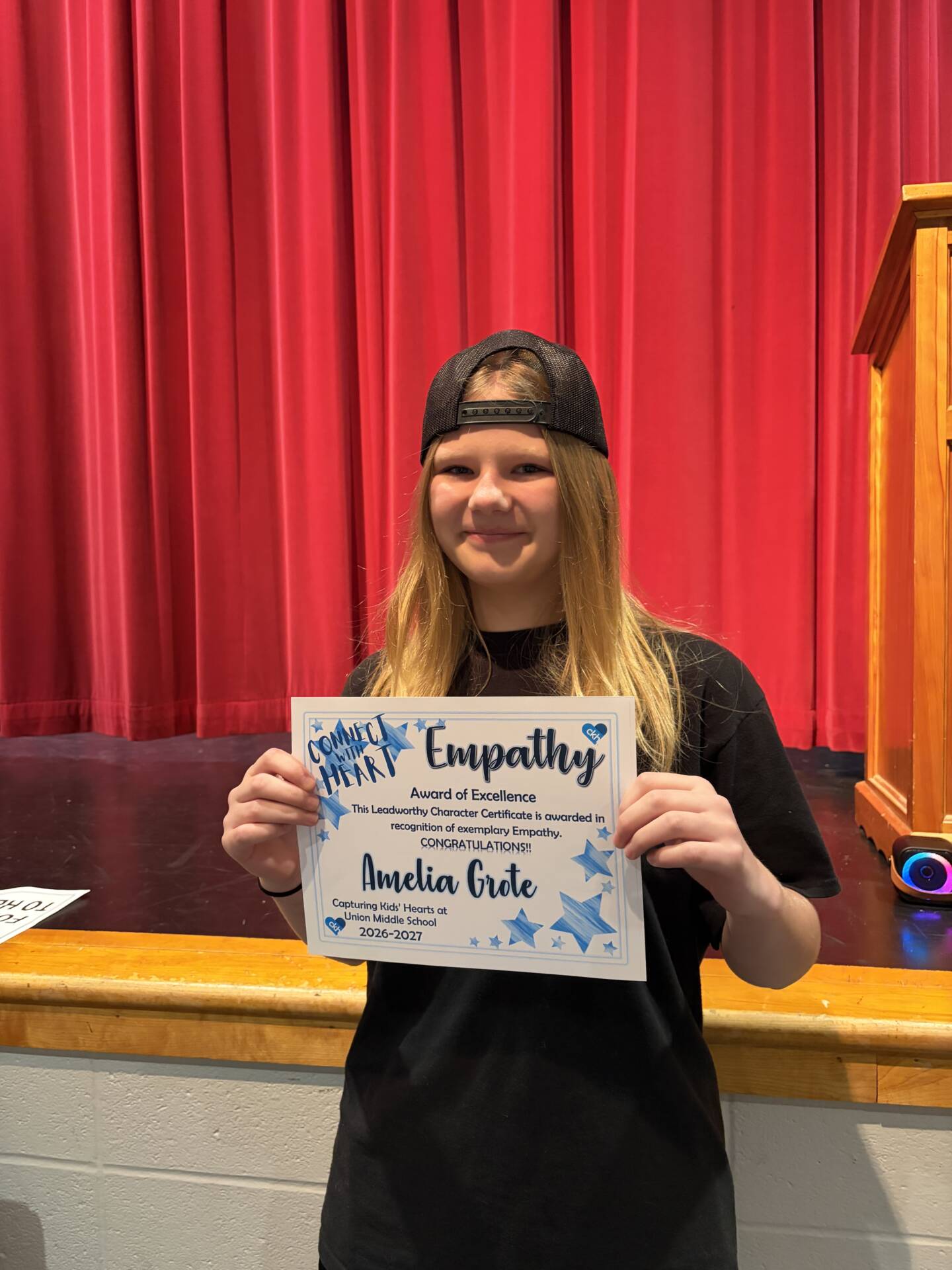 Amelia Grote proudly displays her Empathy Award of Excellence certificate in front of a red stage curtain.
