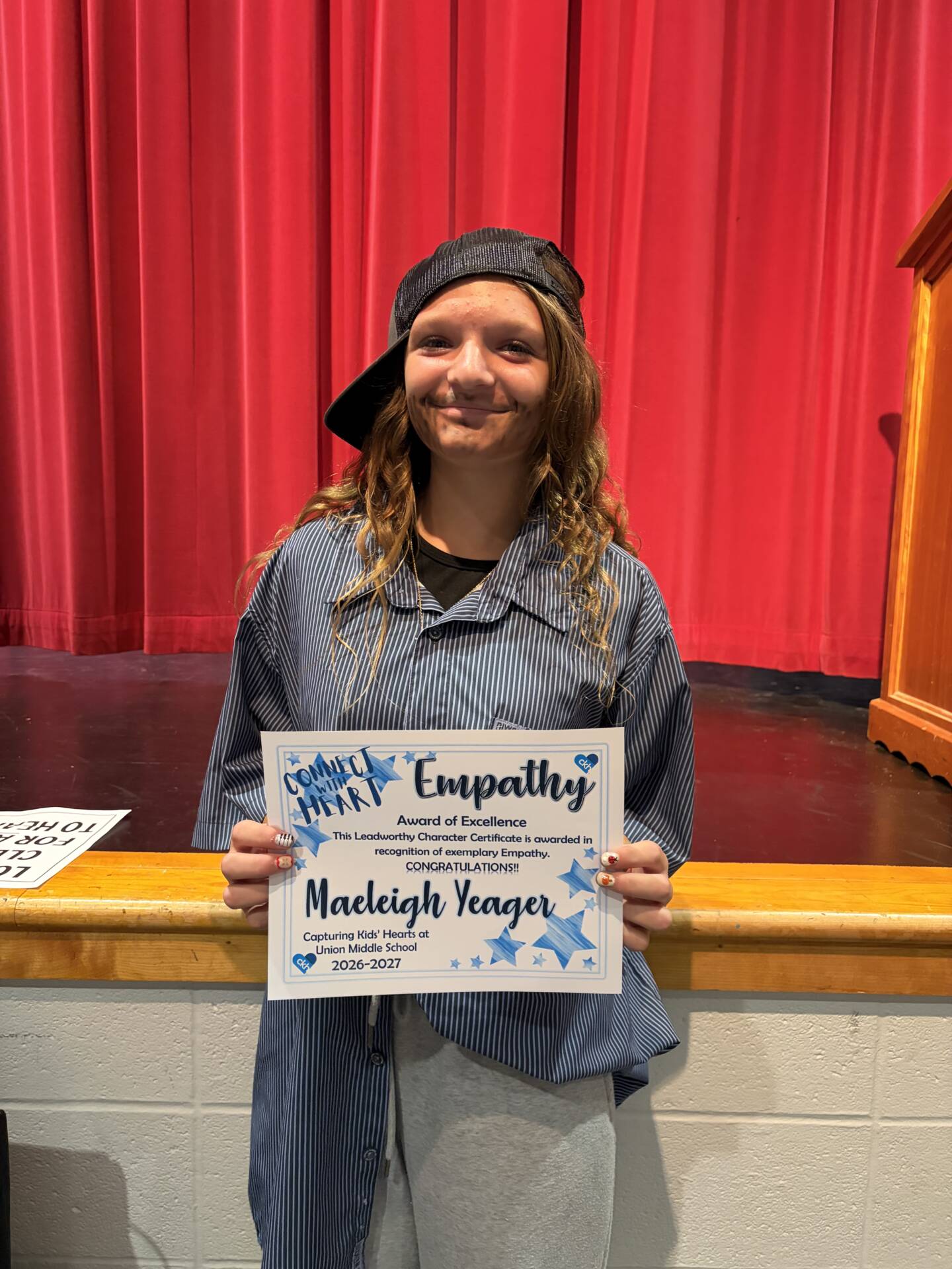 Maeleigh Yeager smiles while holding an Empathy Award of Excellence certificate in front of a red stage curtain.
